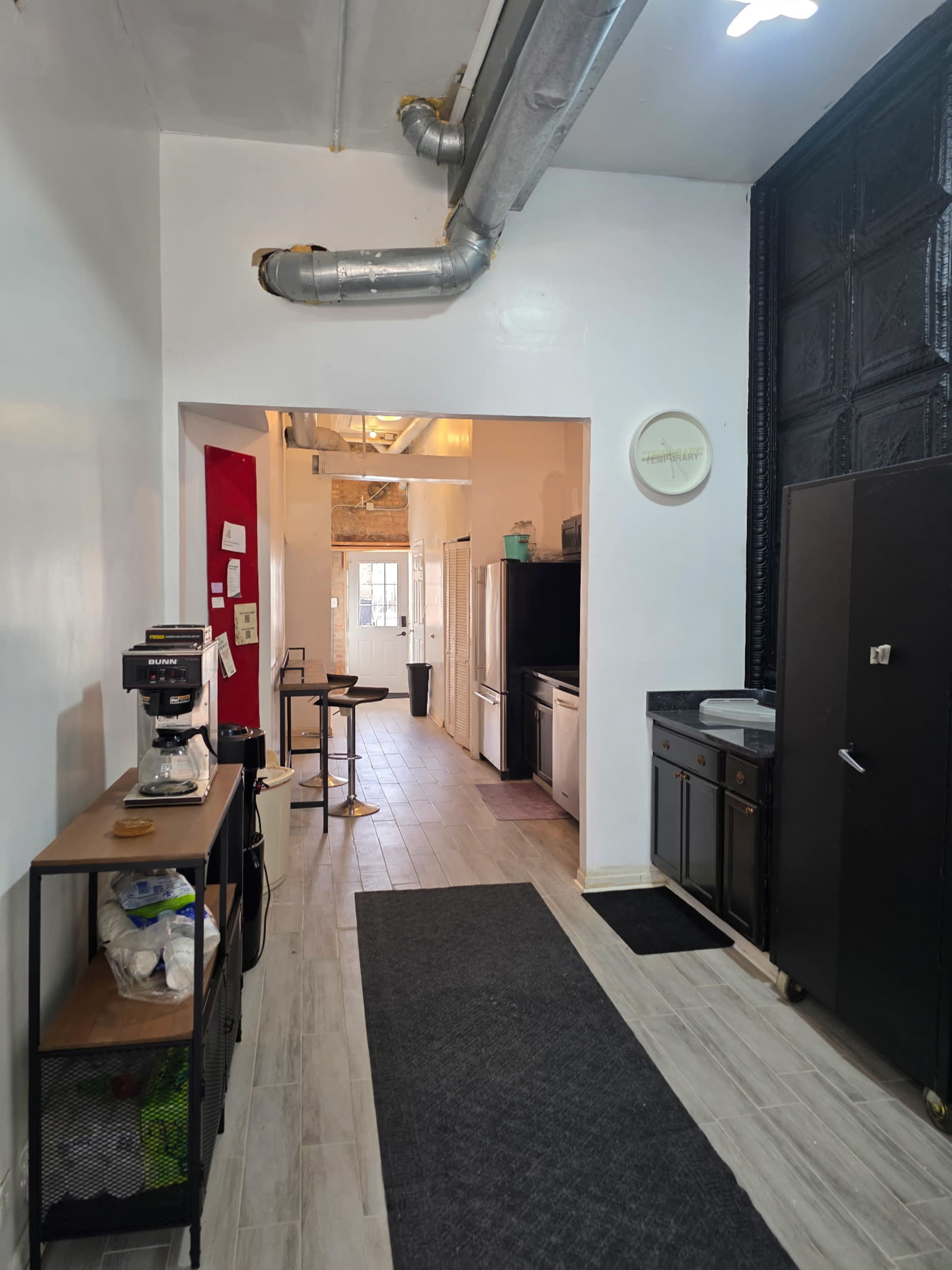 The image shows a narrow kitchen space with stainless steel appliances, a coffee maker on a side table, and a black refrigerator against one wall, leading to another room through an open doorway.