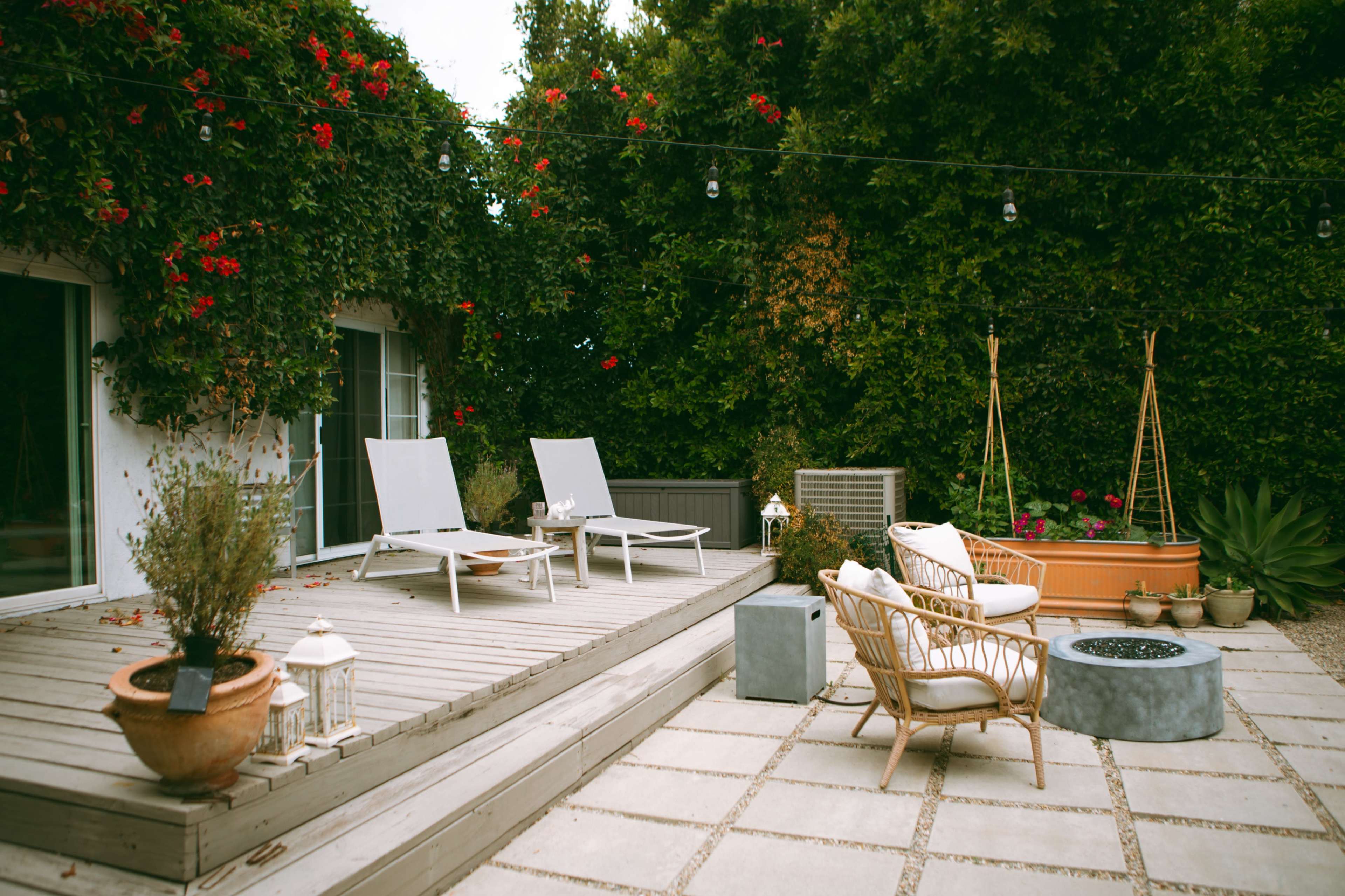 The image shows a patio area with two lounge chairs, a round fire pit, and various potted plants surrounded by vegetated walls.