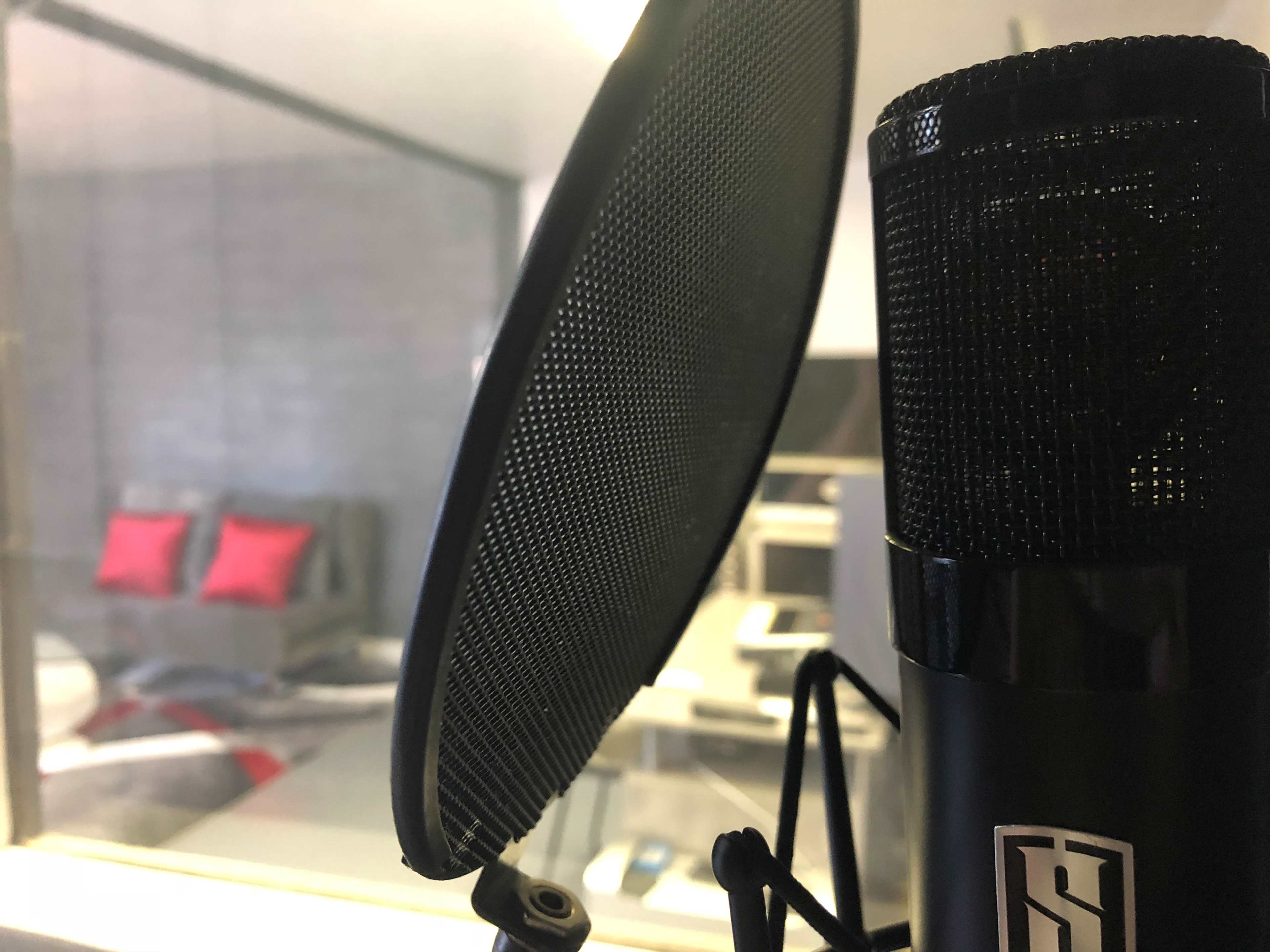 The image shows a microphone with a pop filter in the foreground, while a blurred room with a bed and red pillows is visible in the background.