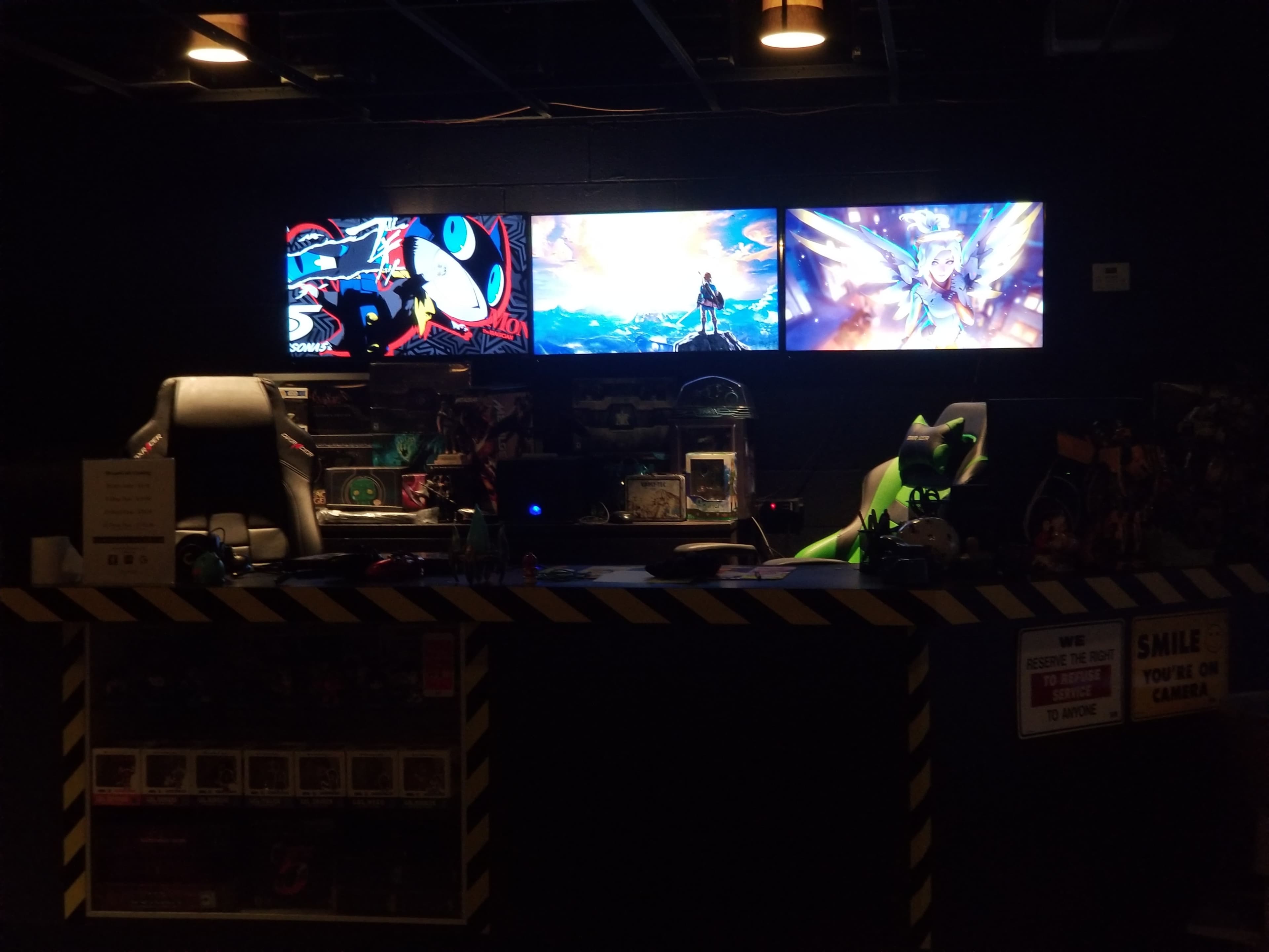 The scene shows a gaming setup with three monitor displays featuring colorful artwork, positioned behind a dark counter with gaming accessories and two gaming chairs.