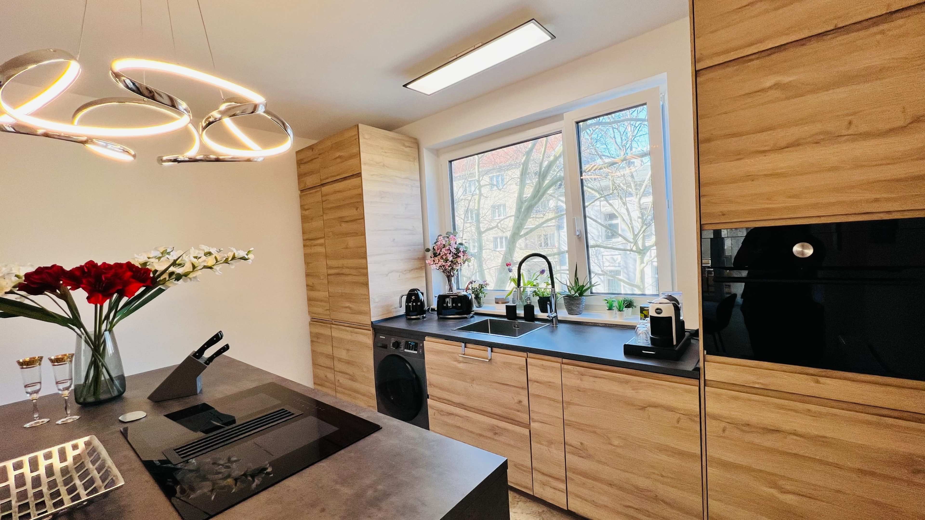 A modern kitchen features wooden cabinetry, a black countertop, a window overlooking a tree-lined street, and a stylish light fixture.