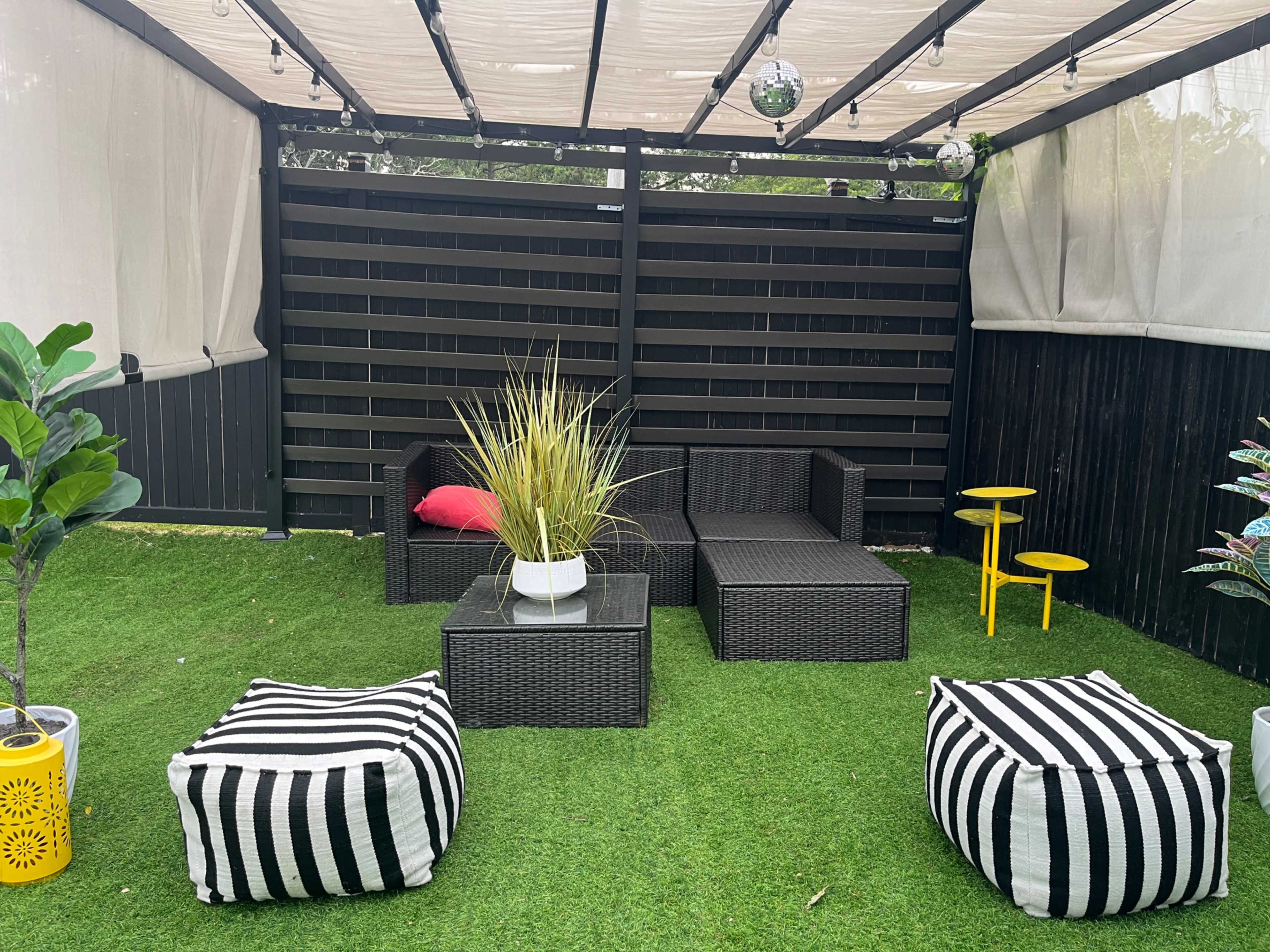 The image shows a shaded outdoor seating area featuring wicker furniture, a decorative plant, and striped poufs on artificial grass.
