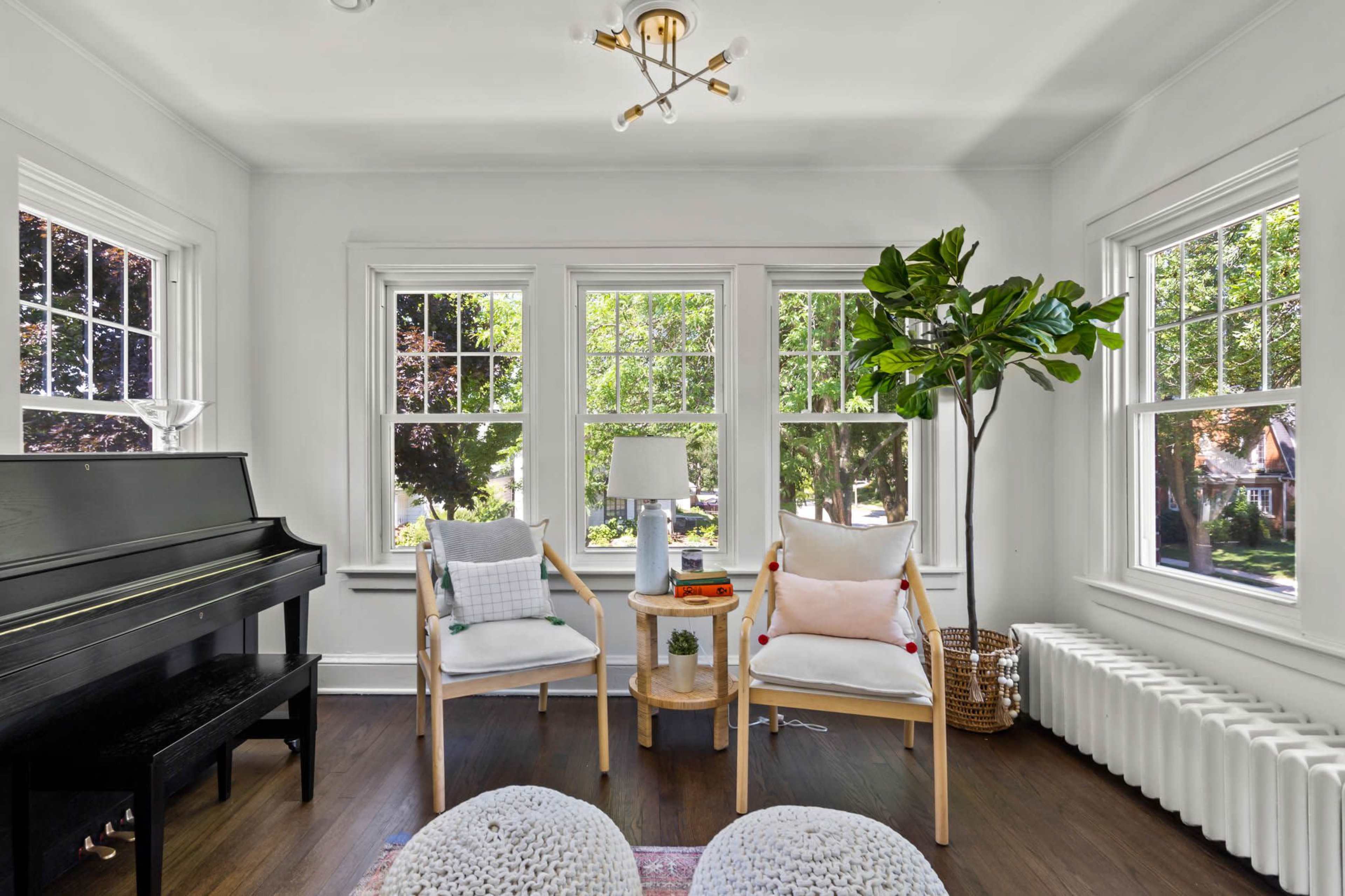 A bright sunroom features two armchairs, a small table with decor, a piano, and large windows overlooking green trees.