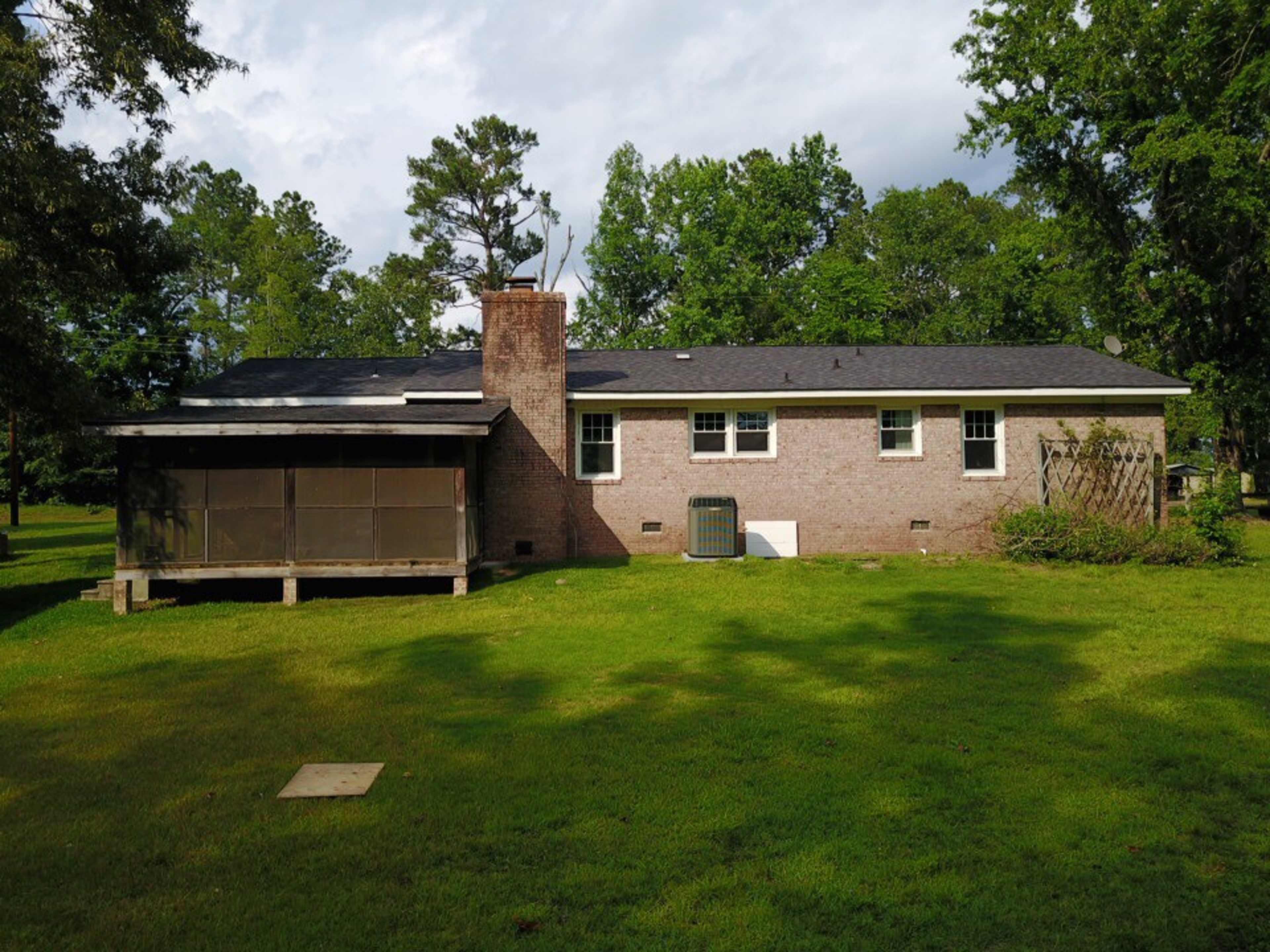 The image shows a single-story brick house with a covered porch and a yard surrounded by trees.