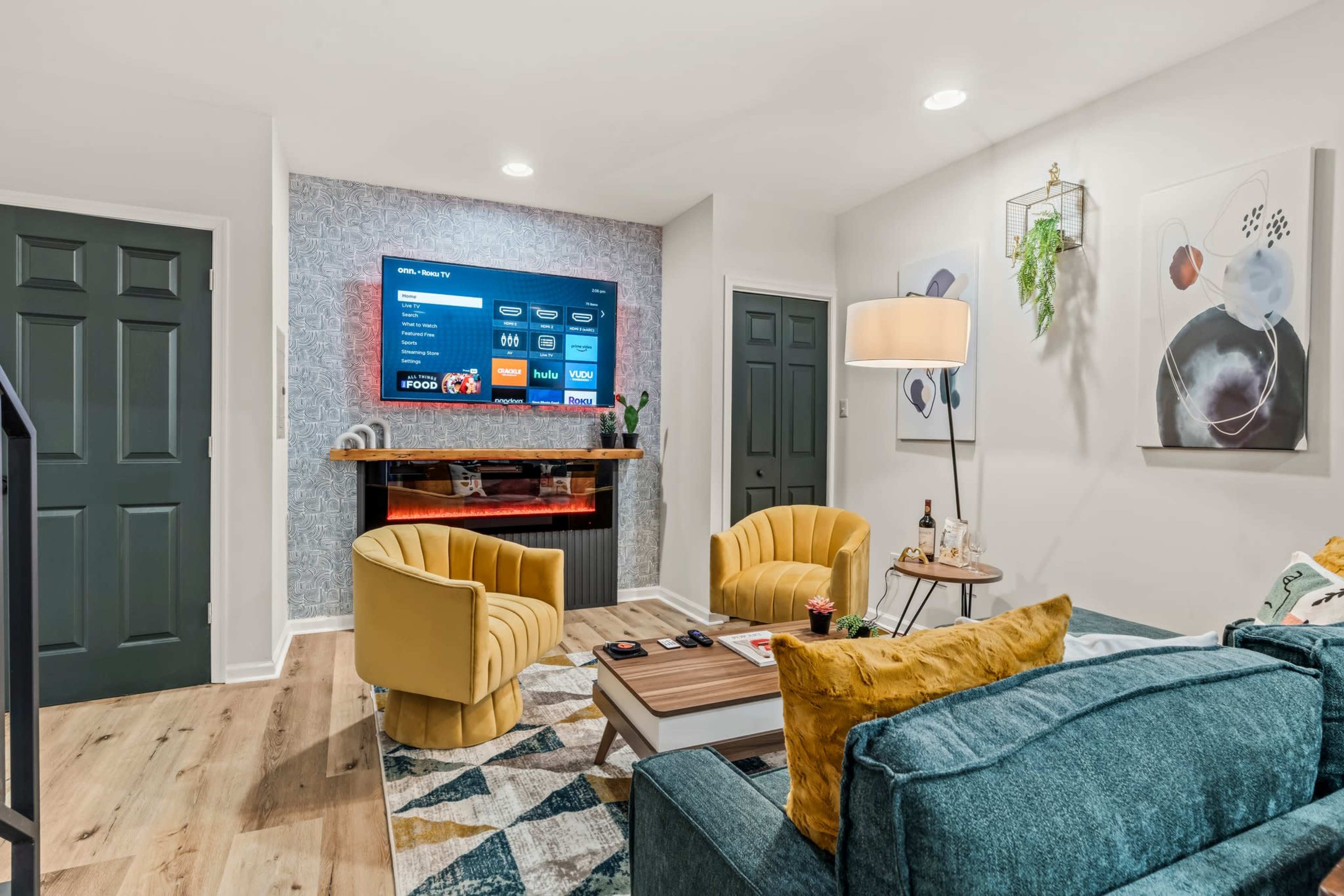 The image shows a modern living room featuring a blue couch, two yellow armchairs, and a wall-mounted television above a wooden shelf.