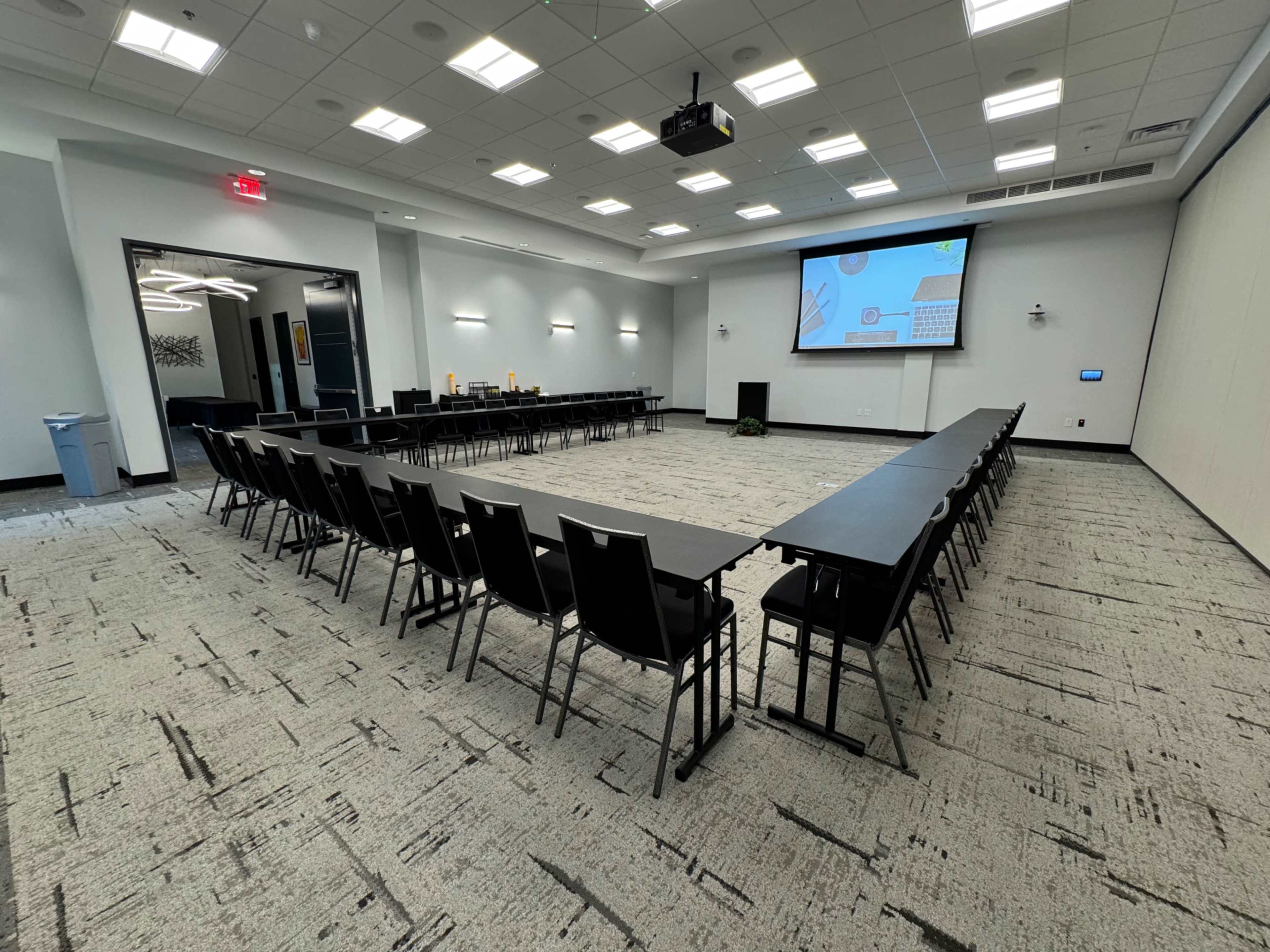 A conference room features a U-shaped arrangement of dark tables and chairs facing a projection screen at one end.