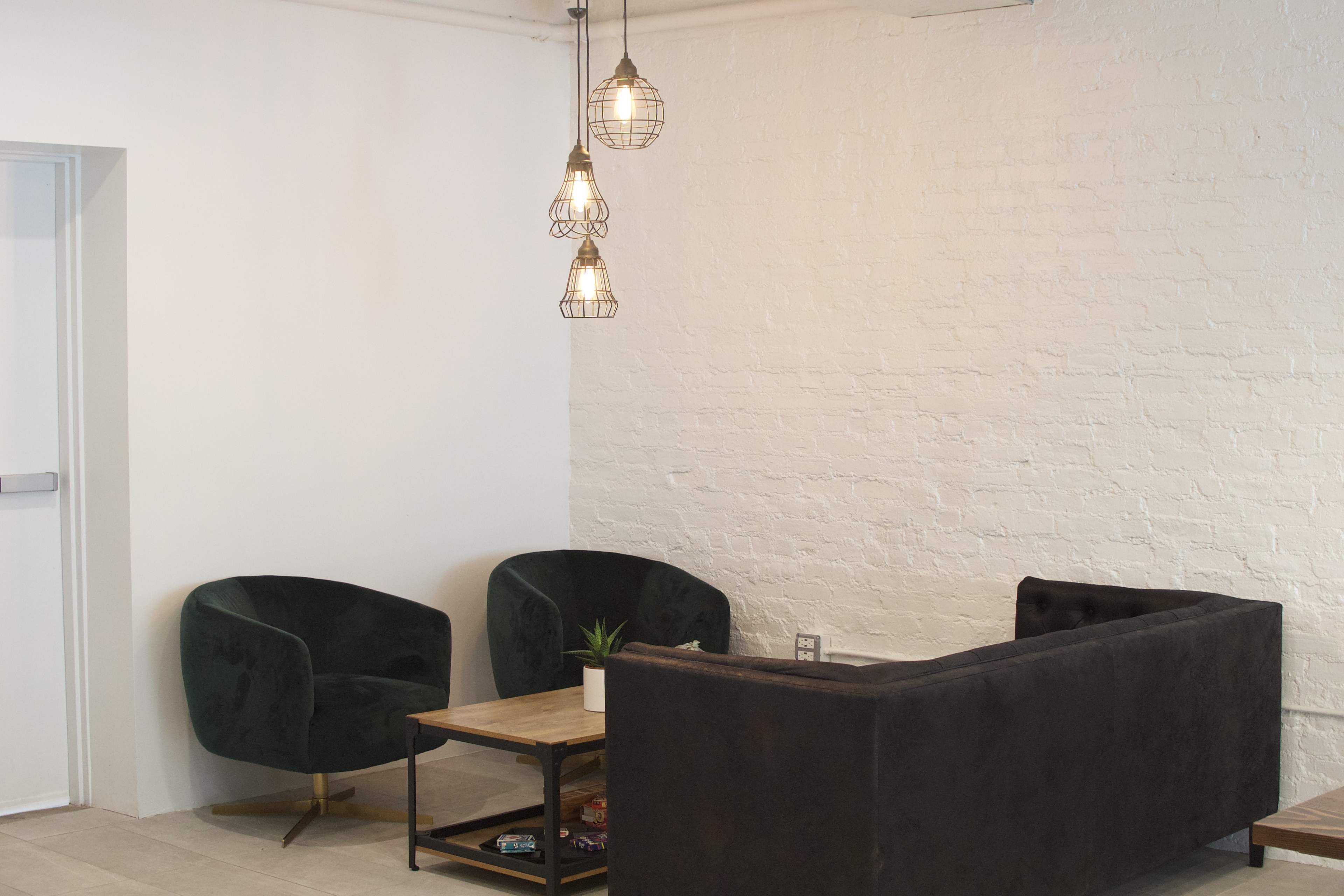 A minimalist interior featuring two green armchairs, a dark sofa, a wooden coffee table, and three pendant lights hanging from the ceiling.