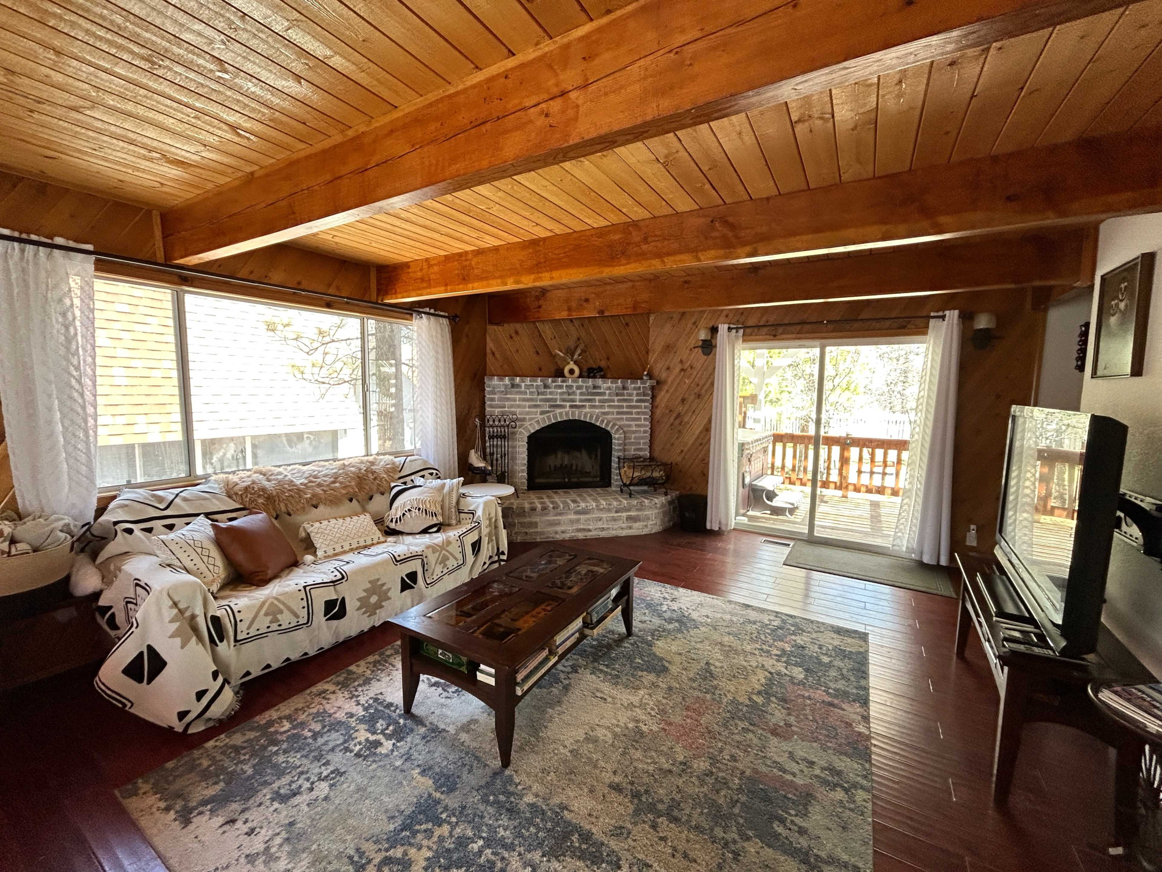 A cozy living room featuring a brick fireplace, wooden beams, a patterned area rug, a sofa with decorative cushions, and large windows that open to a deck.