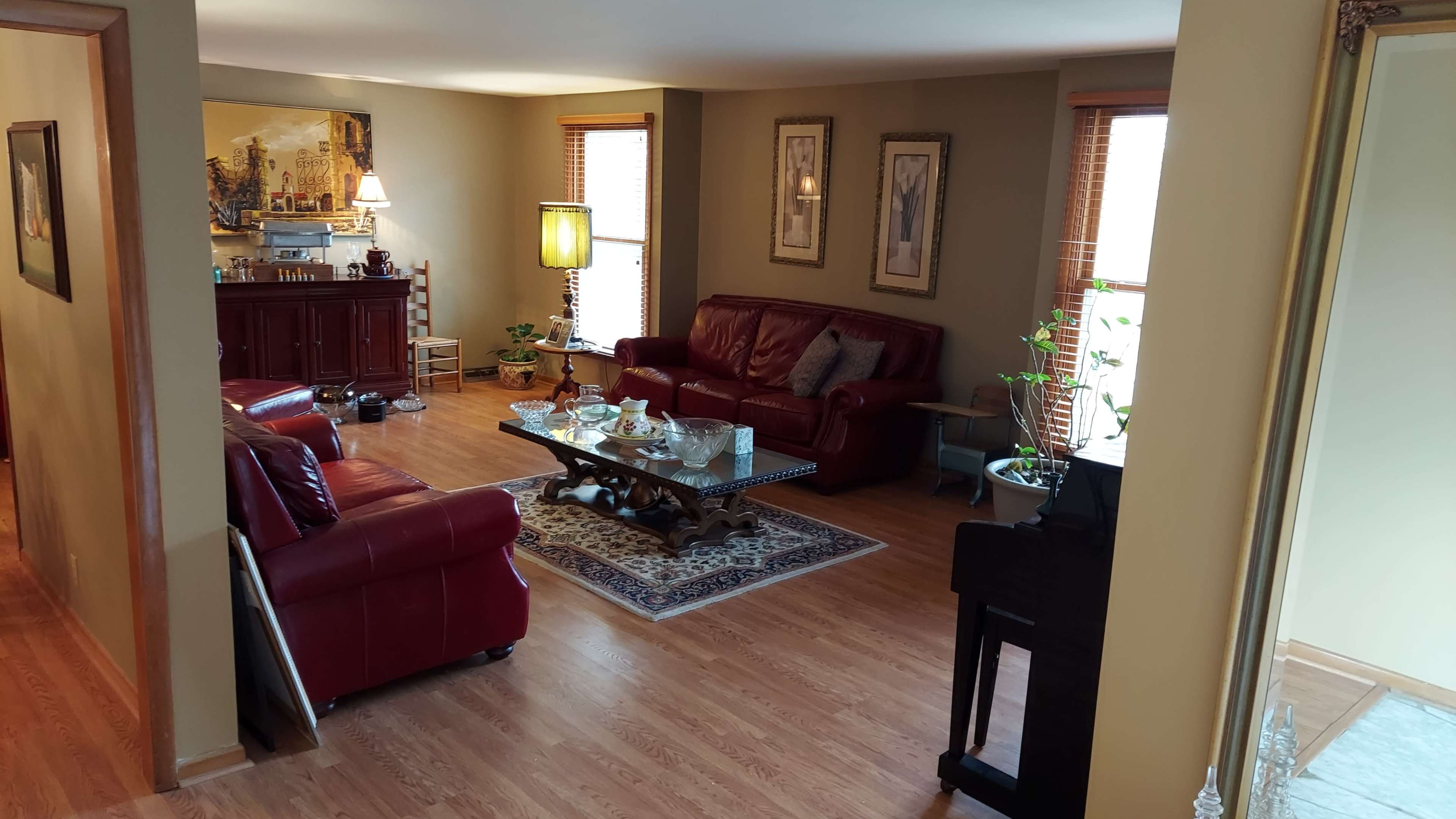 A living room with red leather sofas, a coffee table, and plants, featuring hardwood floors and large windows.