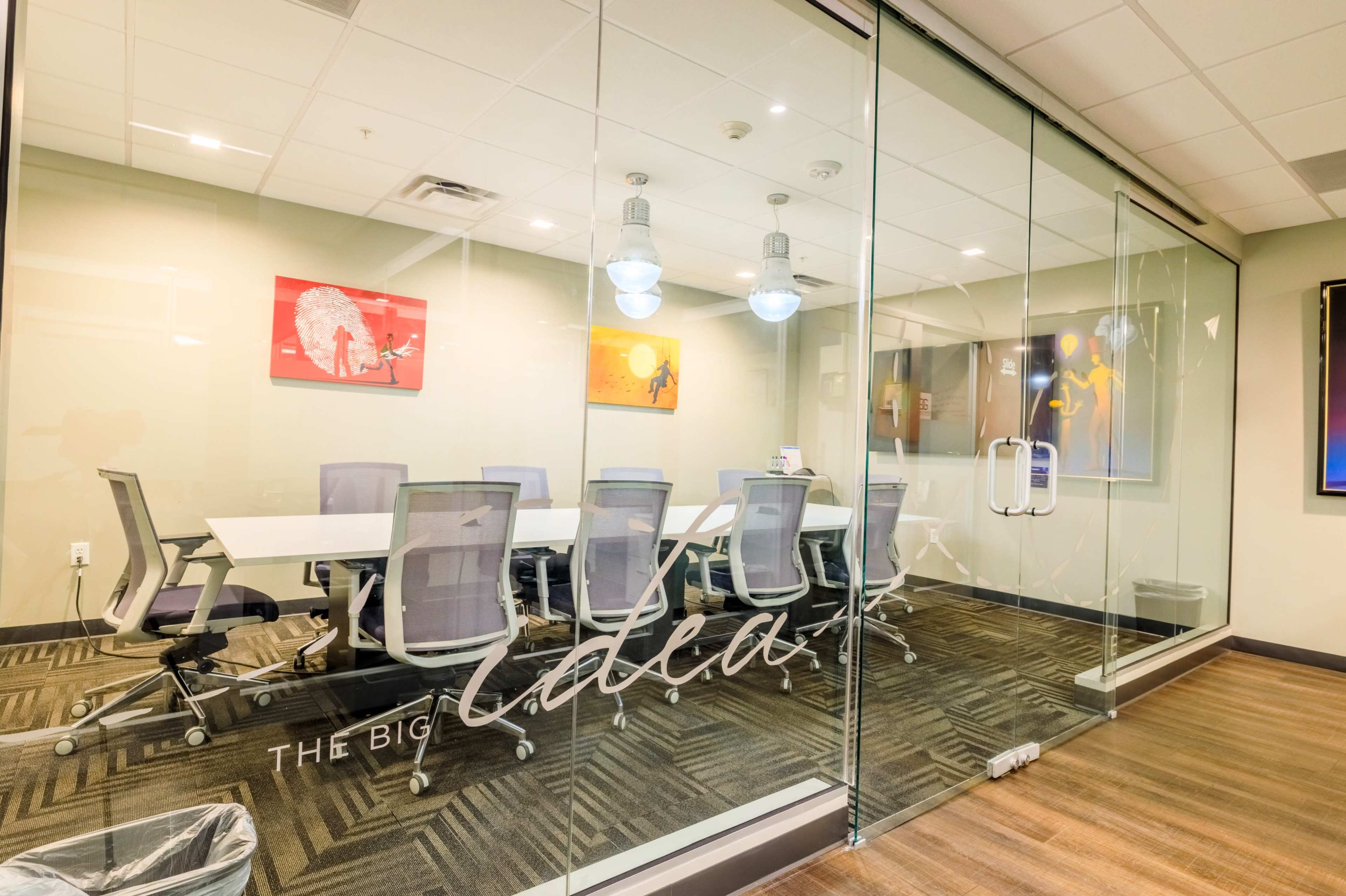 The image shows a modern conference room with a long table surrounded by ergonomic chairs, enclosed by glass walls and featuring vibrant artwork on the walls.