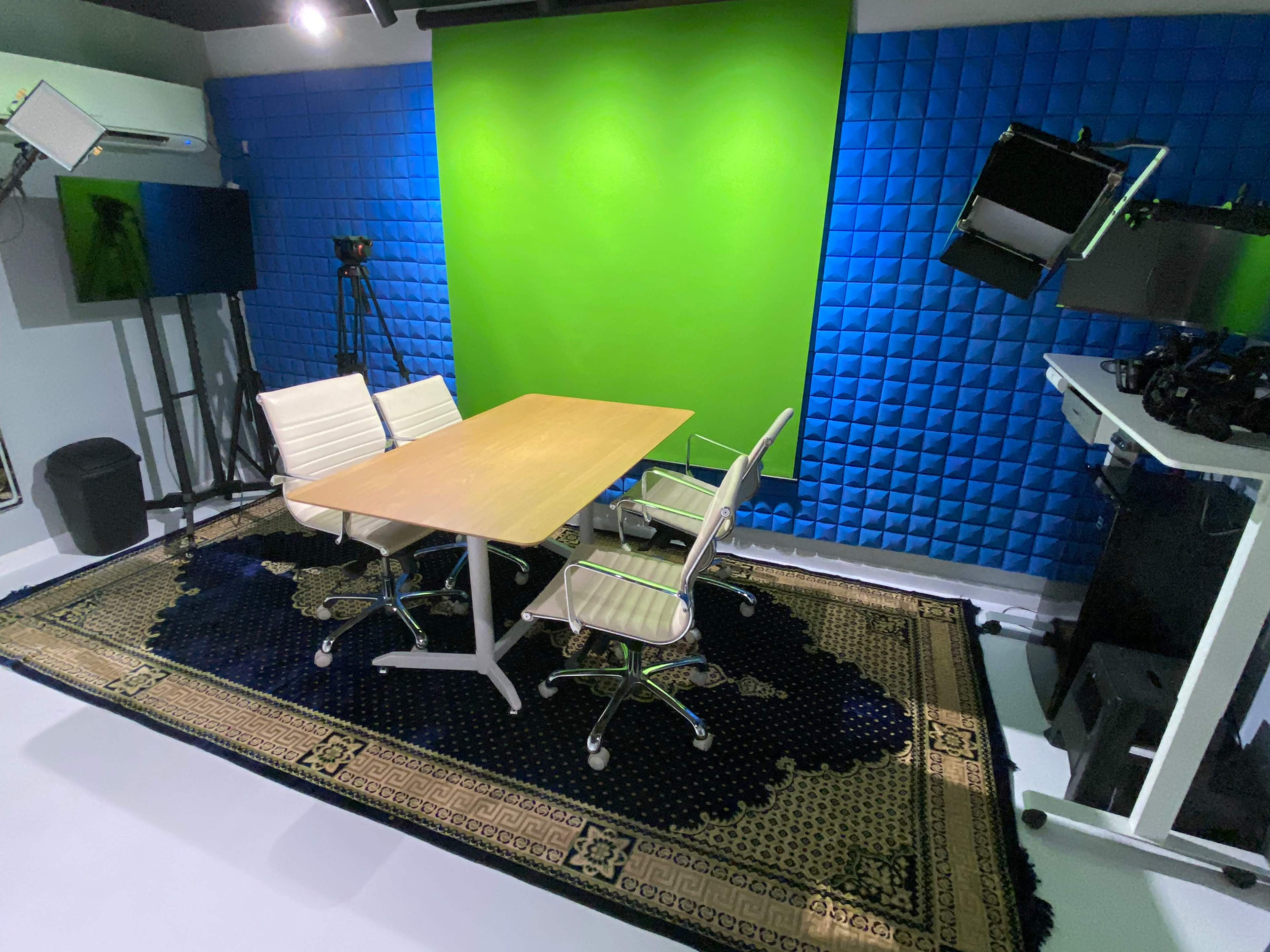 The image shows a recording studio with a green screen background, a wooden table, and three chairs, surrounded by various camera equipment and lighting.