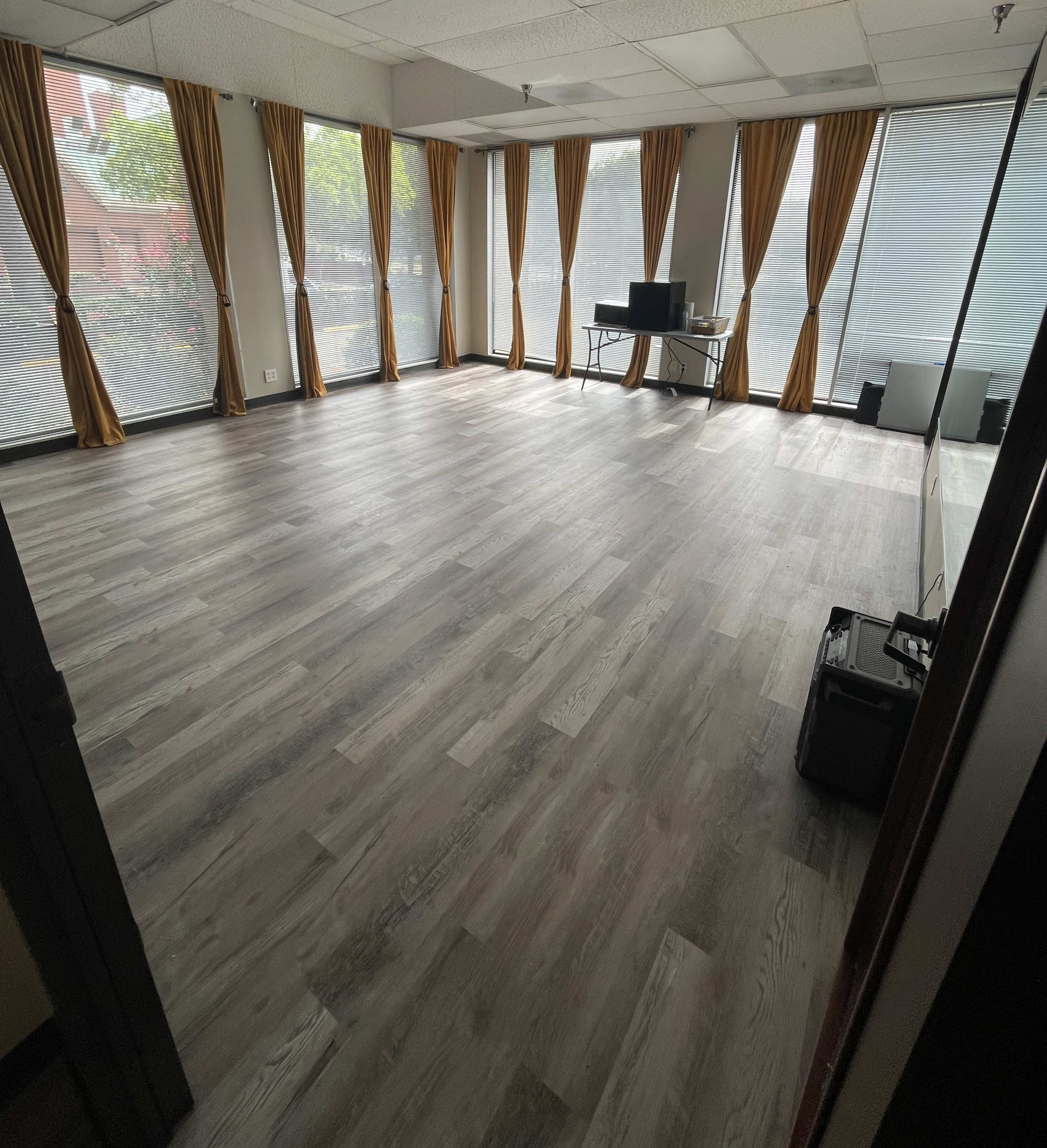 The image shows a vacant room with wooden flooring, large windows covered by sheer curtains, and a desk with a computer set up against one wall.