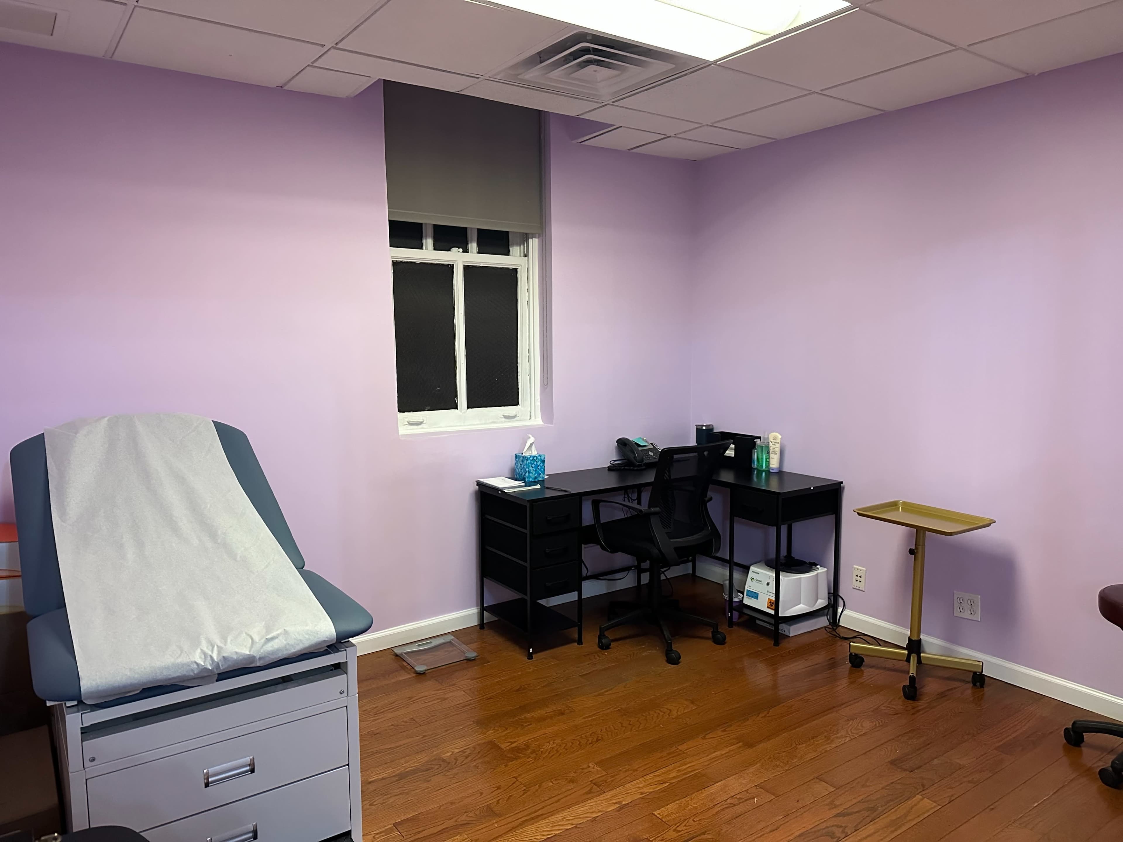 The image shows a small medical examination room with a patient examination table, a black desk and chair, and a rolling cart in a space with lavender-colored walls and hardwood flooring.