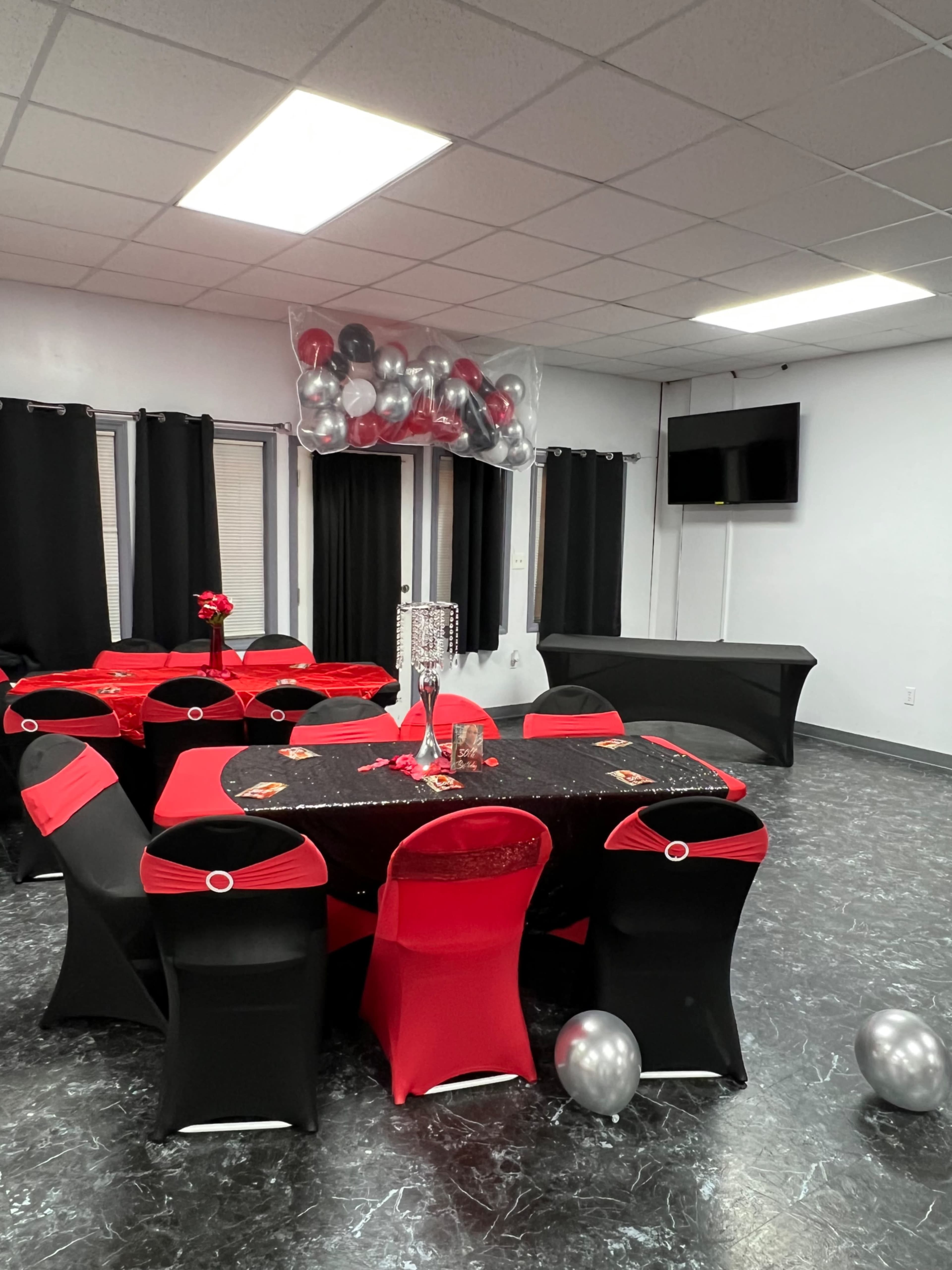 A banquet hall is set up with tables covered in black and red linens, decorated with silver balloons, and red flowers, while black curtains hang in the background.