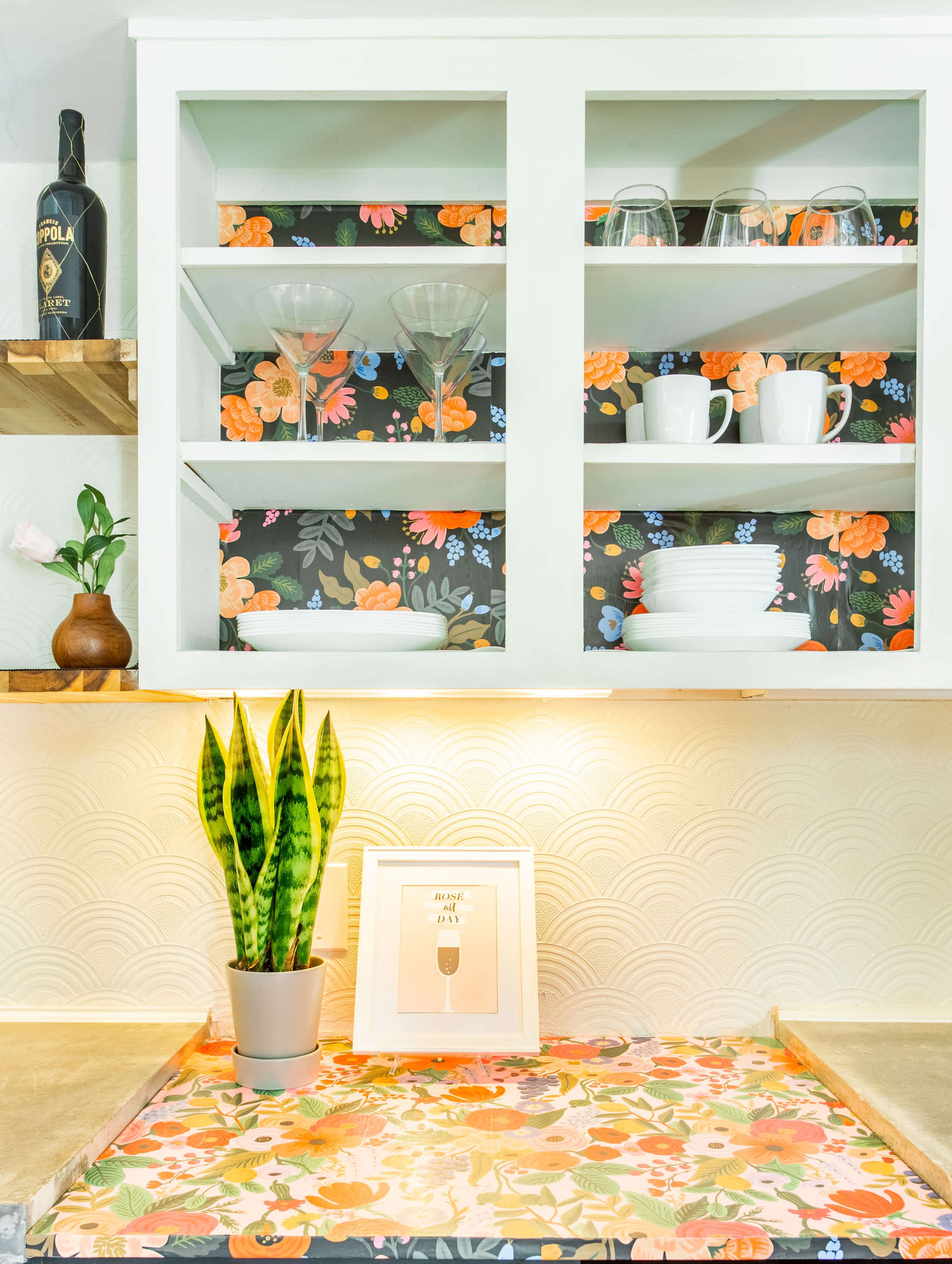 A modern kitchen features open shelves filled with glassware and dishware, a decorative plant on the counter, and floral wallpaper backdrop.