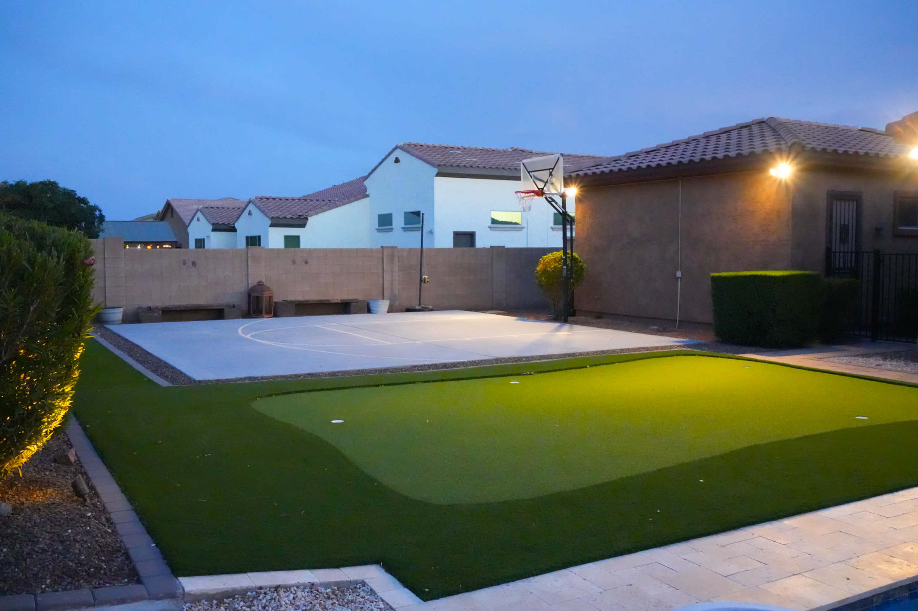 The image shows a residential backyard featuring a basketball court and a putting green, surrounded by low walls and nearby houses at dusk.