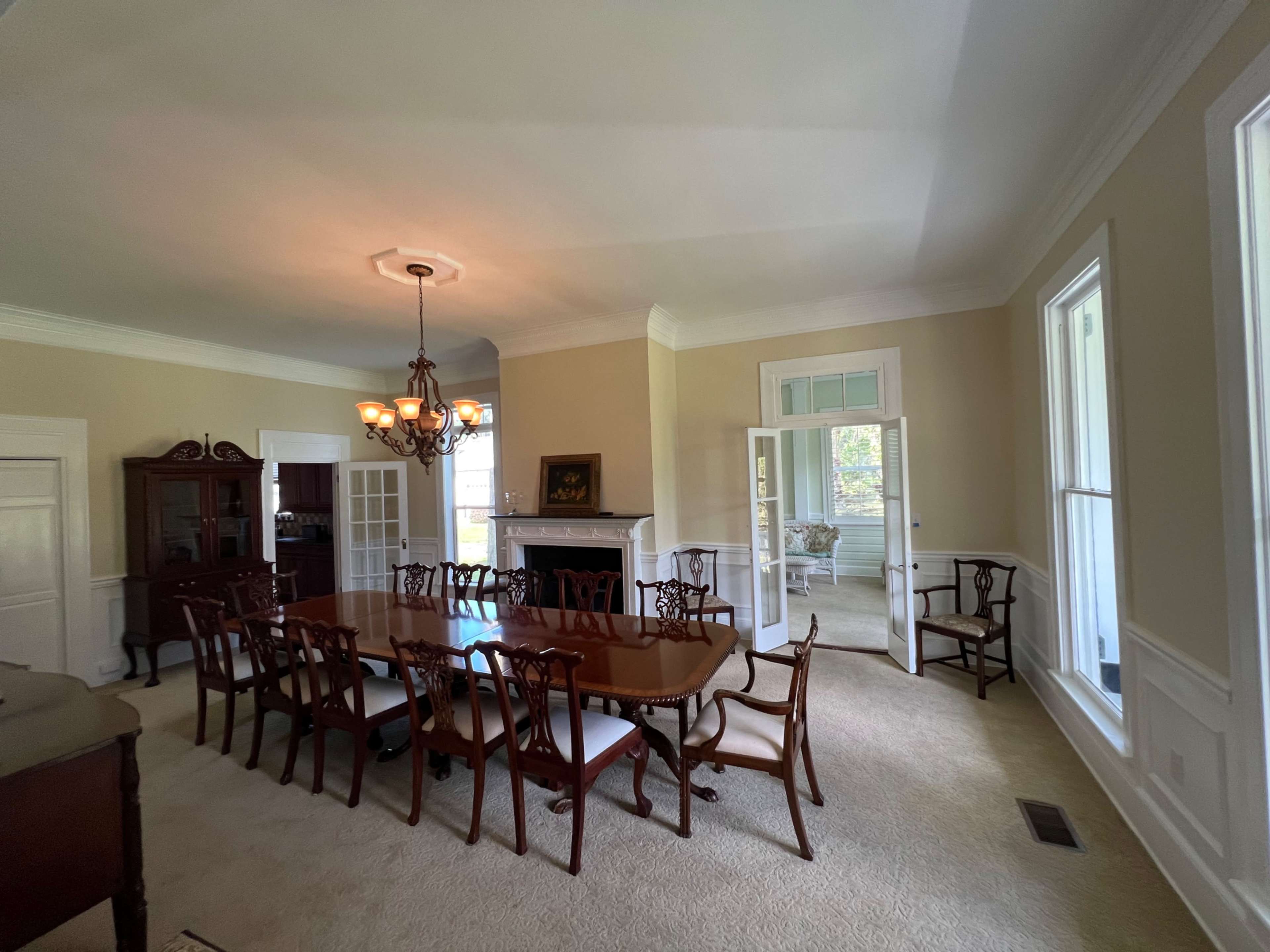The image shows a spacious dining room featuring a large, rectangular wooden table surrounded by chairs, with a chandelier and a fireplace visible in the background.