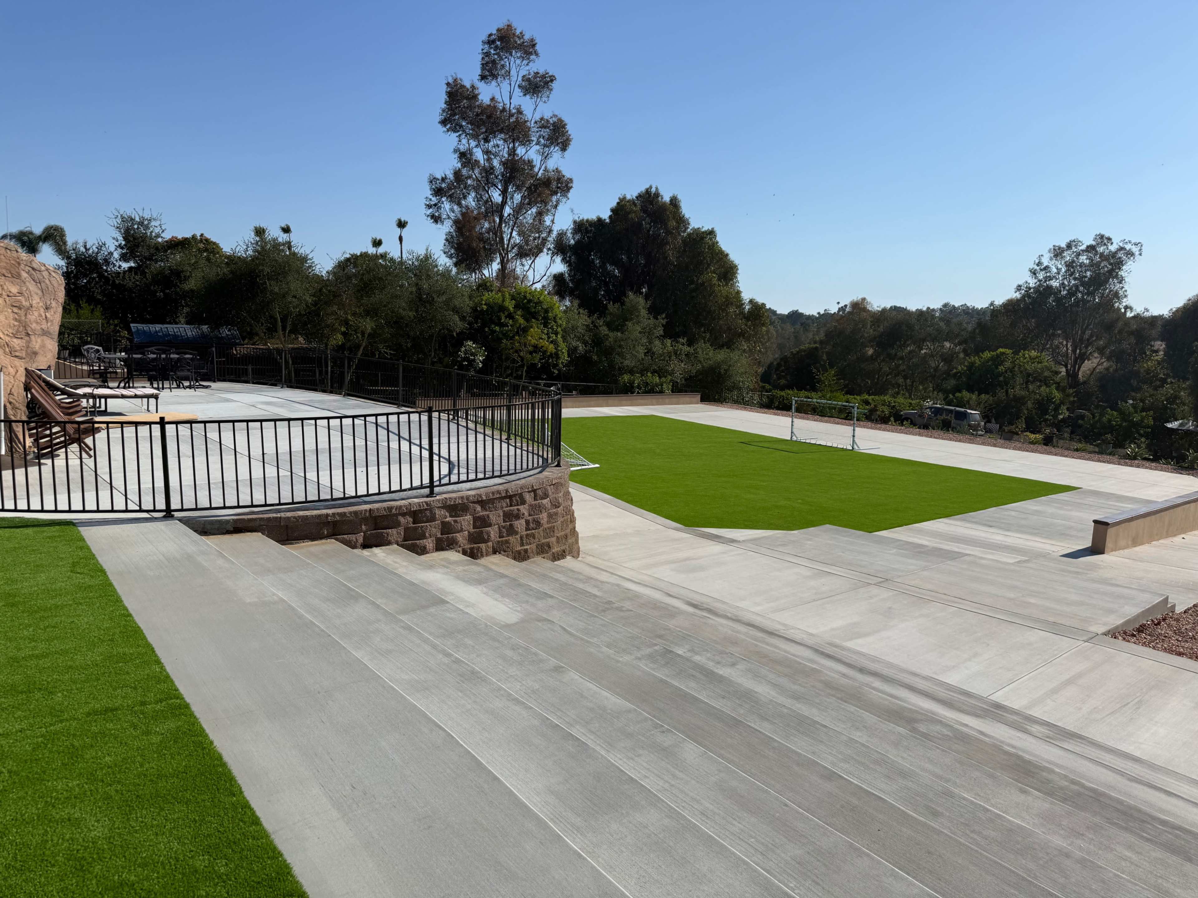 The image shows a landscaped outdoor area featuring a combination of concrete surfaces and green artificial grass, with seating along the perimeter against a backdrop of trees and hills.