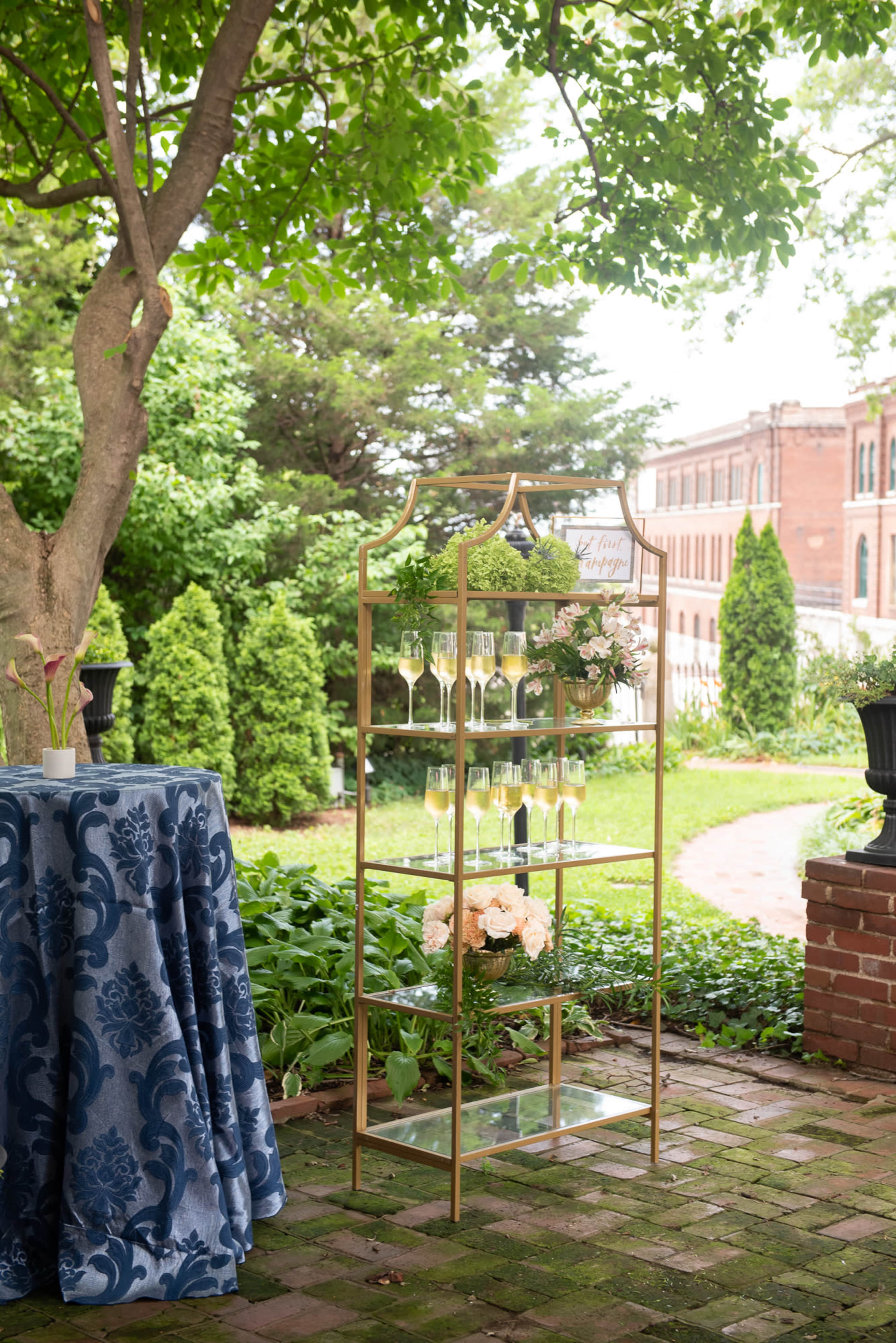 A decorative metal shelving unit displays champagne flutes alongside floral arrangements in an outdoor garden setting.