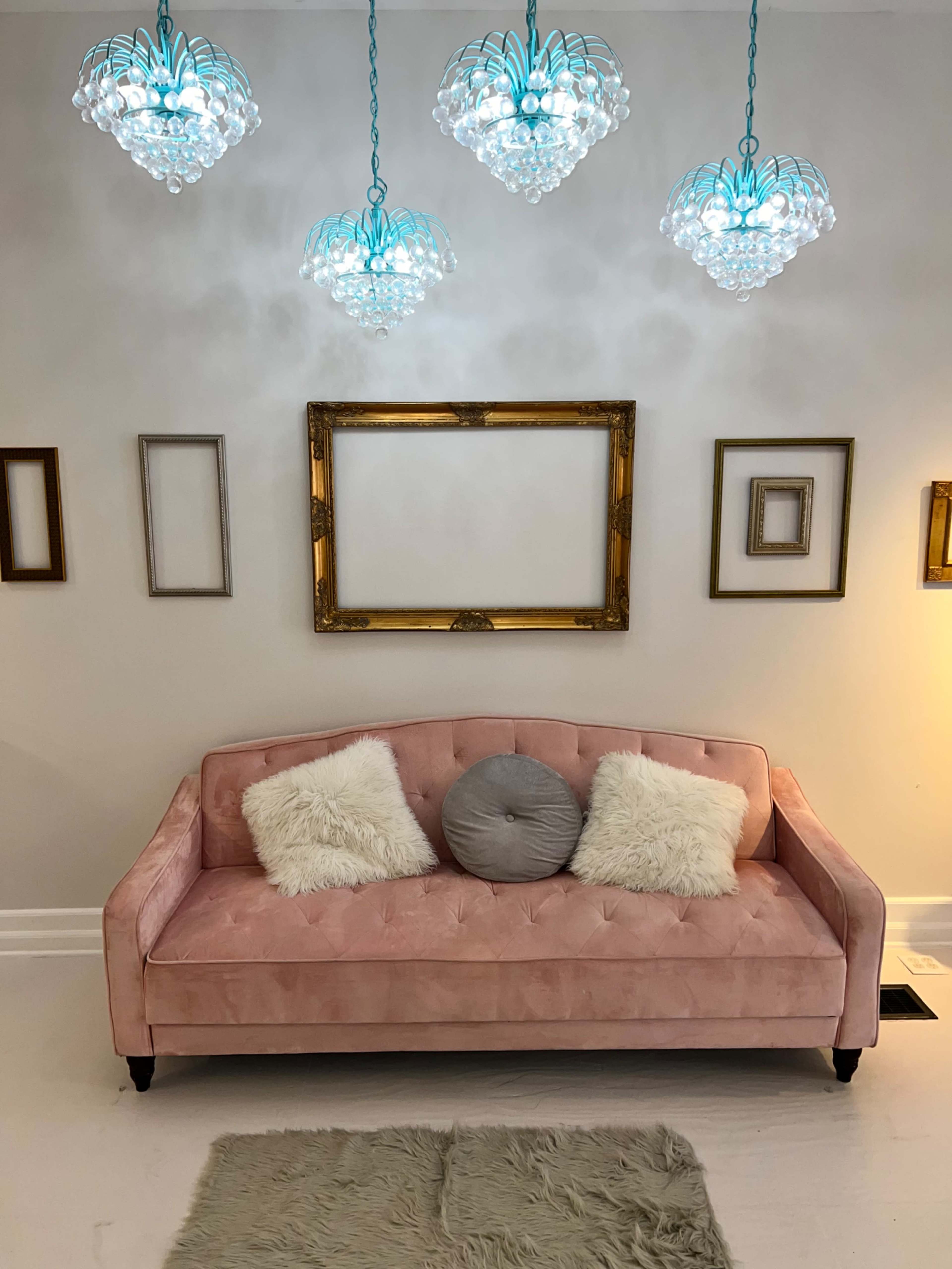 A pink sofa with decorative pillows is positioned against a light-colored wall adorned with various empty frames and three pendant lights above.