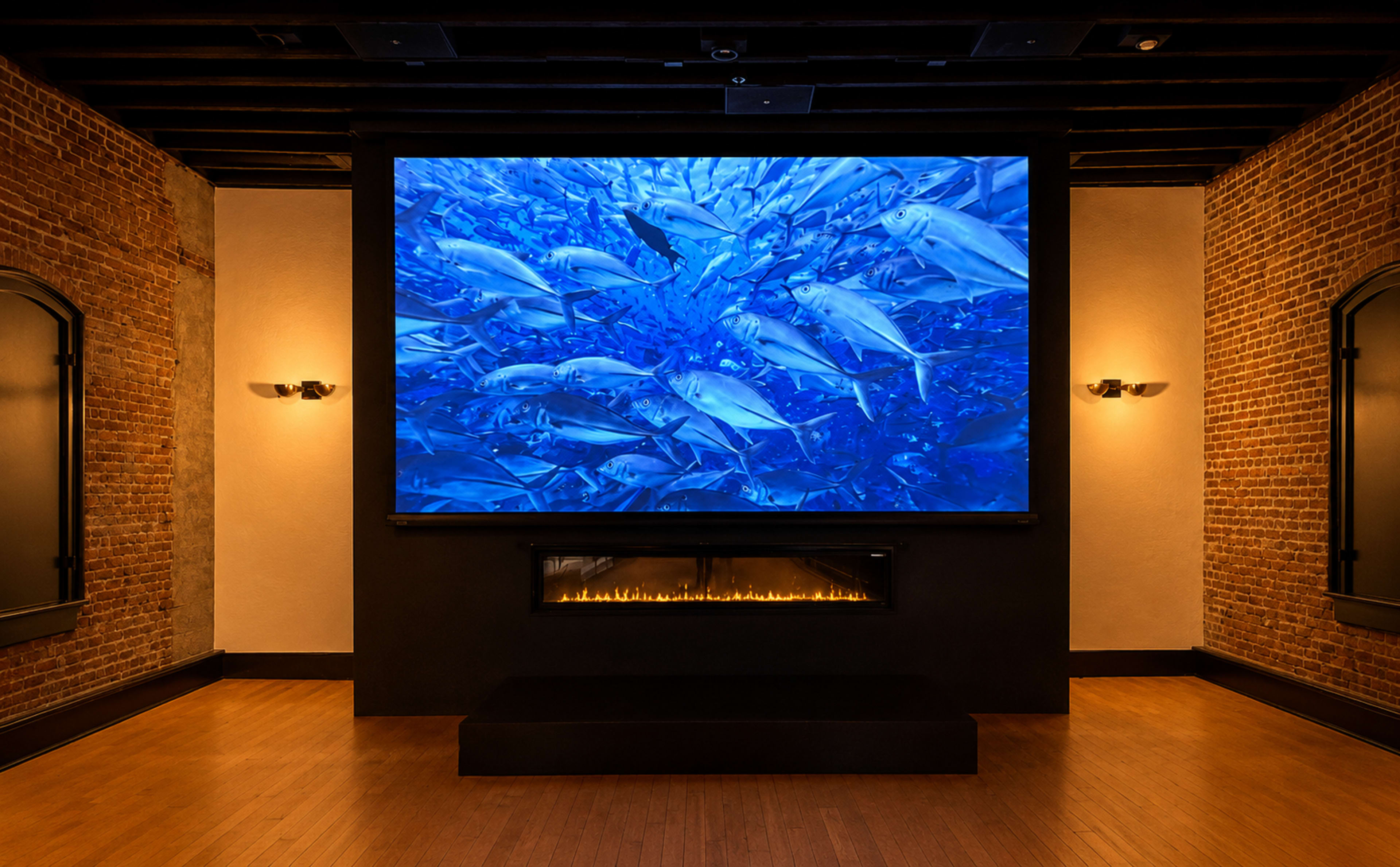The image shows a modern room with exposed brick walls, featuring a large screen displaying a school of fish and a fireplace underneath.