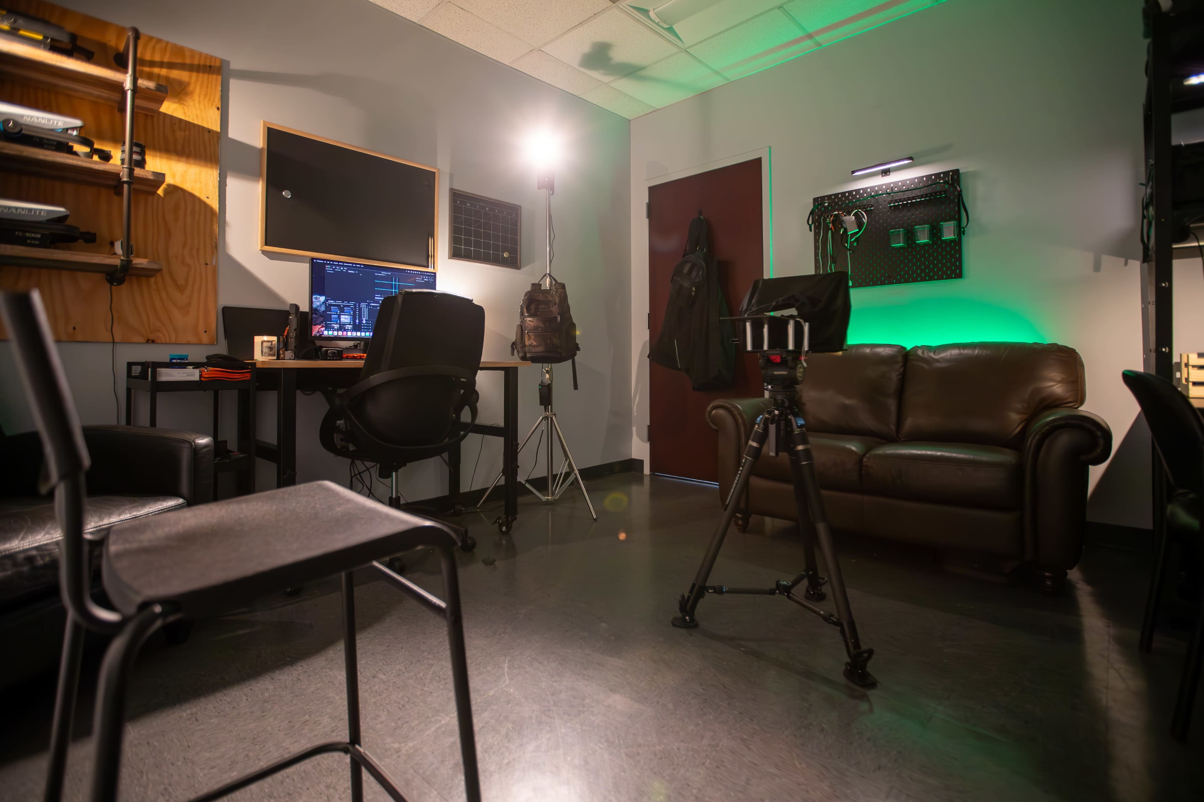 The image shows a small studio space with a brown leather couch, a desk with a computer setup, a tripod, and lighting equipment against a wall.
