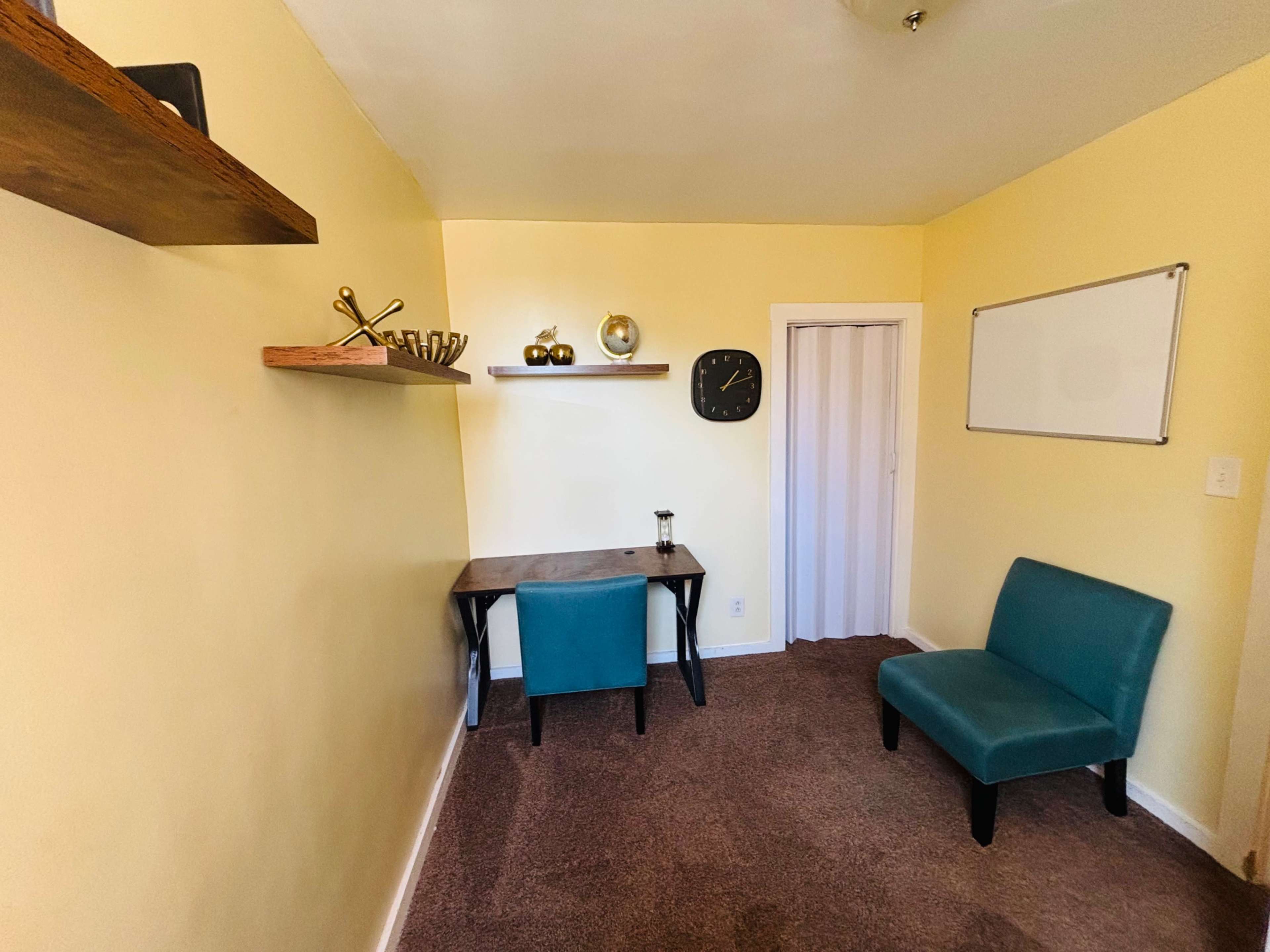 The image shows a small room with light yellow walls, a wooden desk, a teal chair, a wall clock, and a whiteboard on the wall.
