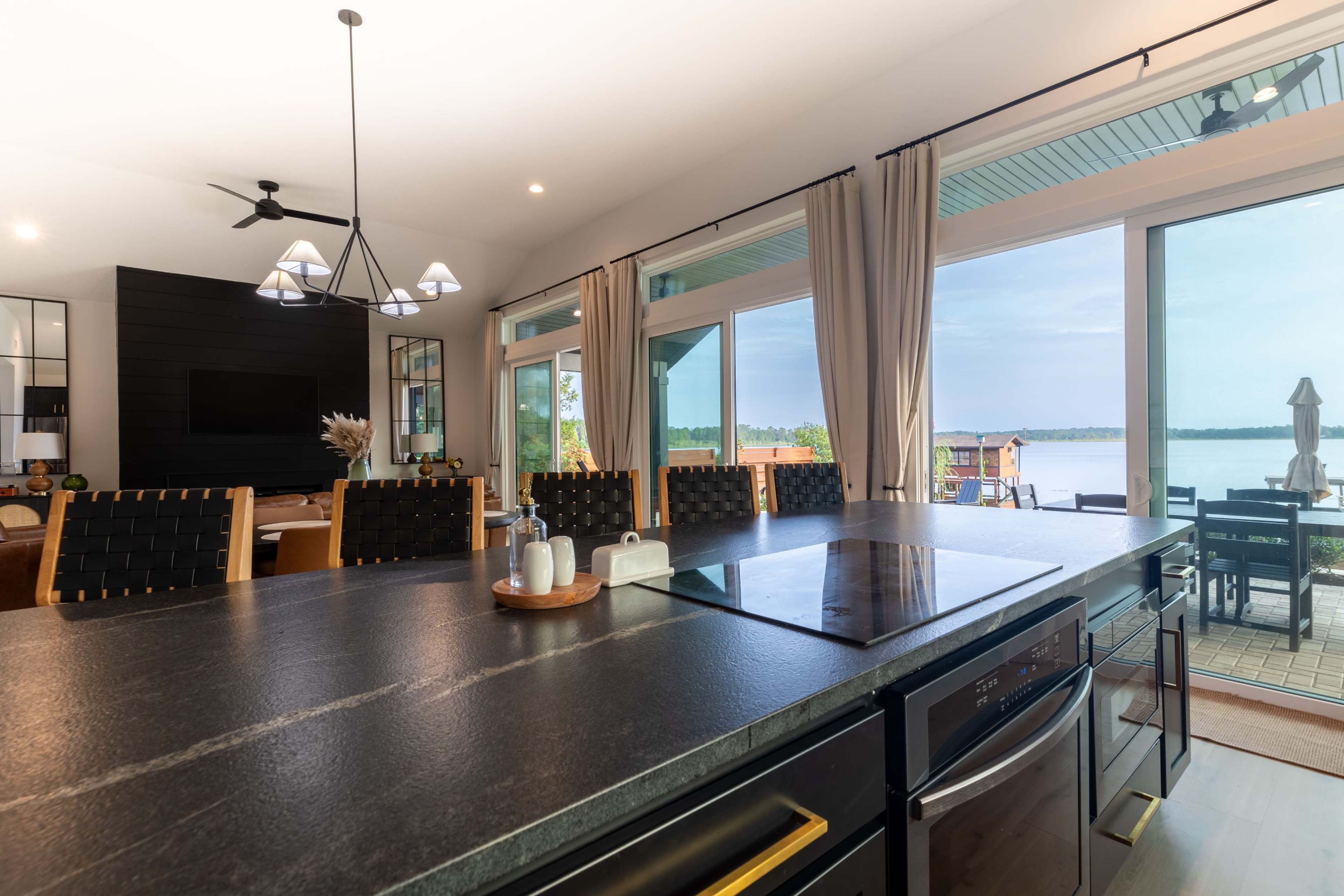 The kitchen features a dark countertop with a cooking surface, overlooking large glass doors that lead to a deck by the water.