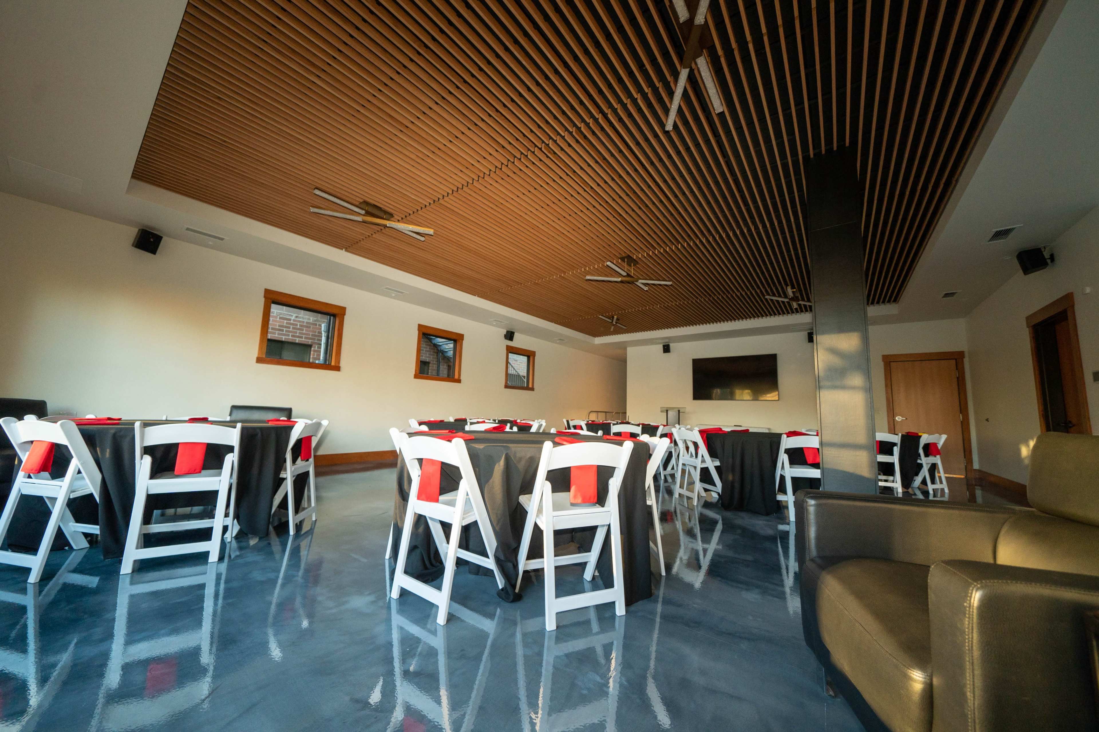 A modern event space with rows of tables covered in black linens and red napkins, set beneath a wooden ceiling with ceiling fans.