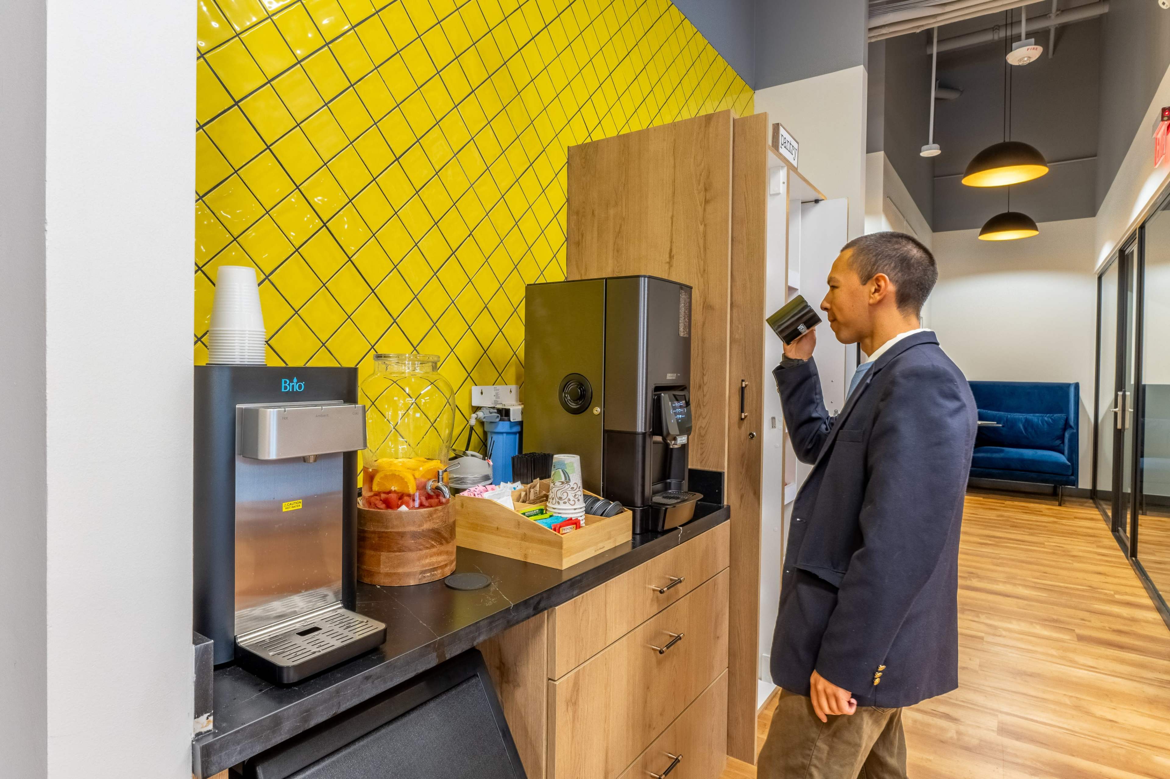 A man in a blazer drinks from a cup near a coffee machine in a modern kitchen with yellow tiled walls and a wooden countertop.