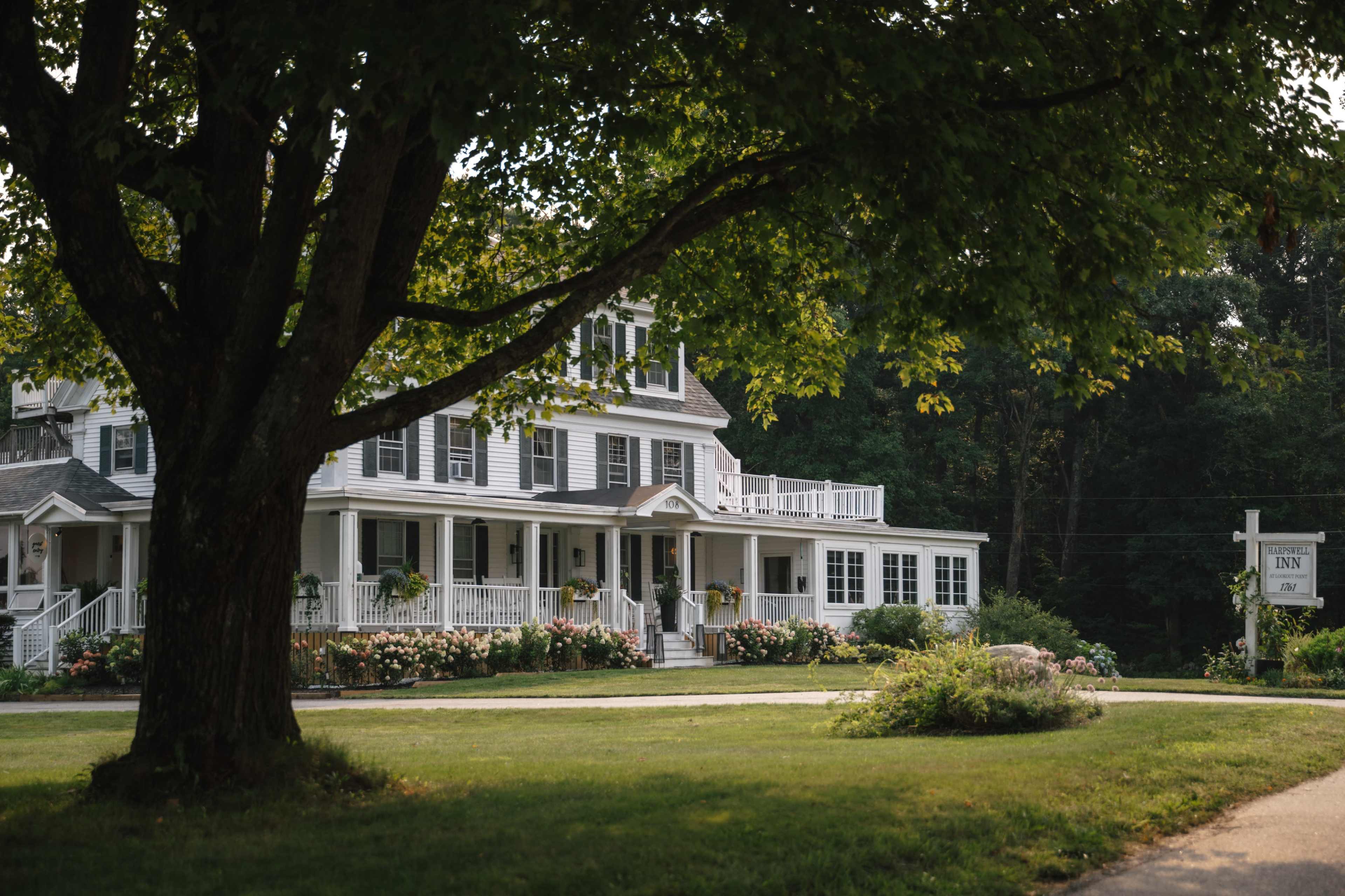 A large, white inn with multiple stories and a wide porch sits under a tree, surrounded by a grassy area and blooming flowers.