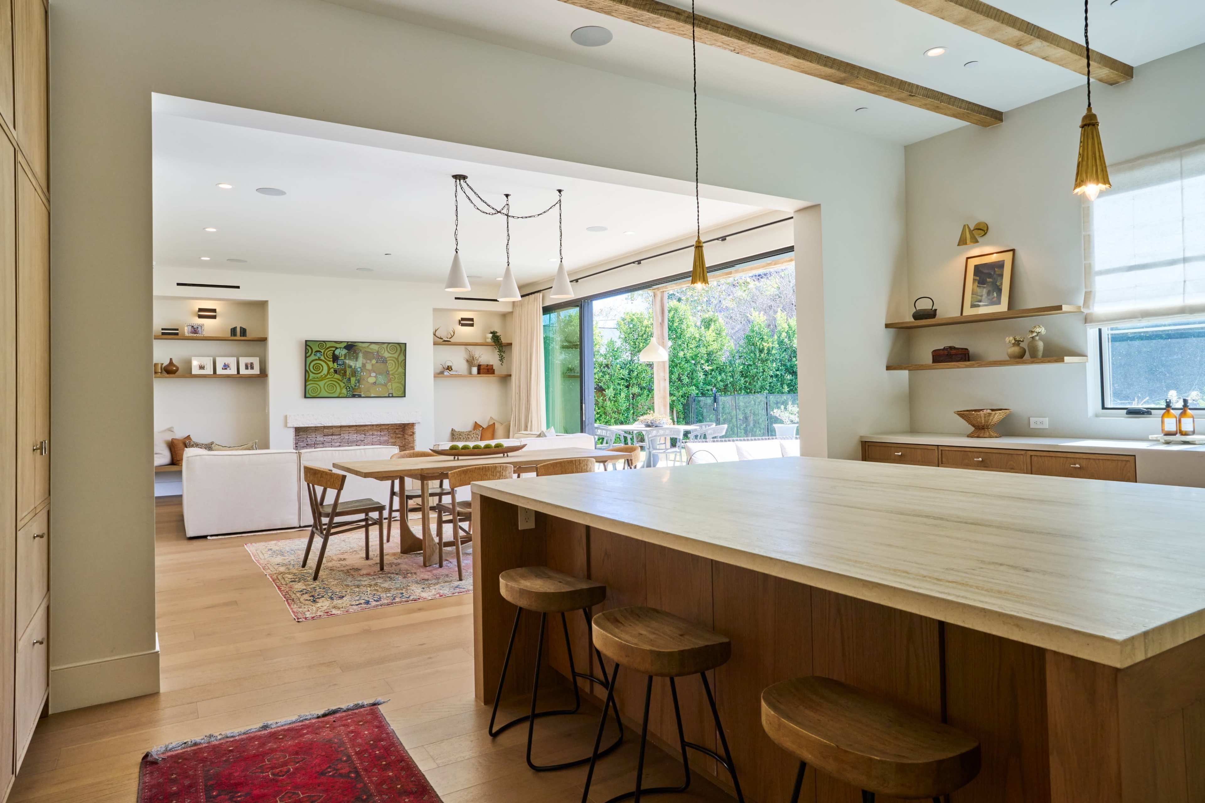 The image shows a modern kitchen with a large wooden island and open access to a light-filled living area, featuring a sliding glass door leading to an outdoor patio.