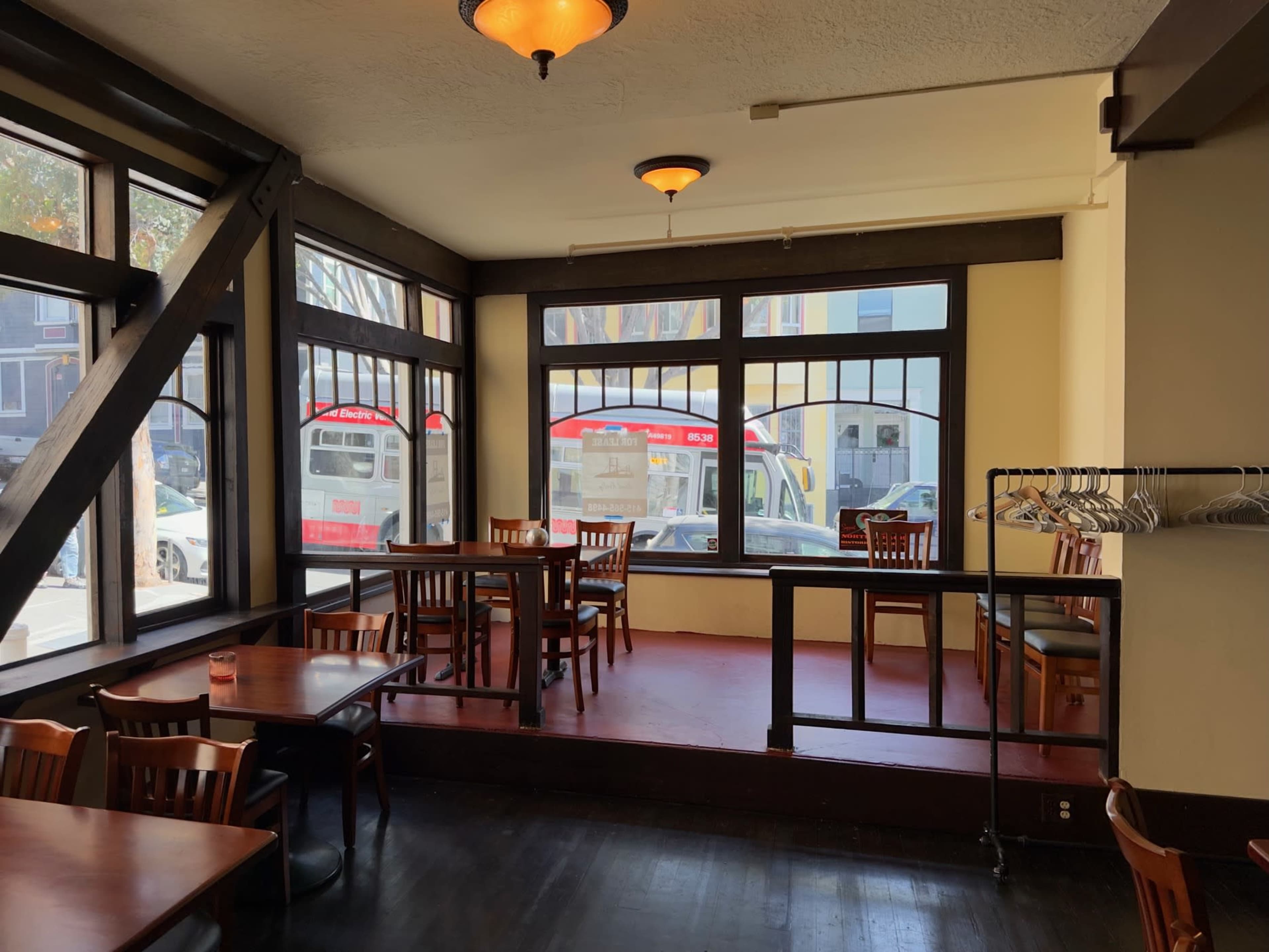 The image shows a cozy restaurant interior with wooden tables and chairs, featuring large windows that provide a view of the street outside.