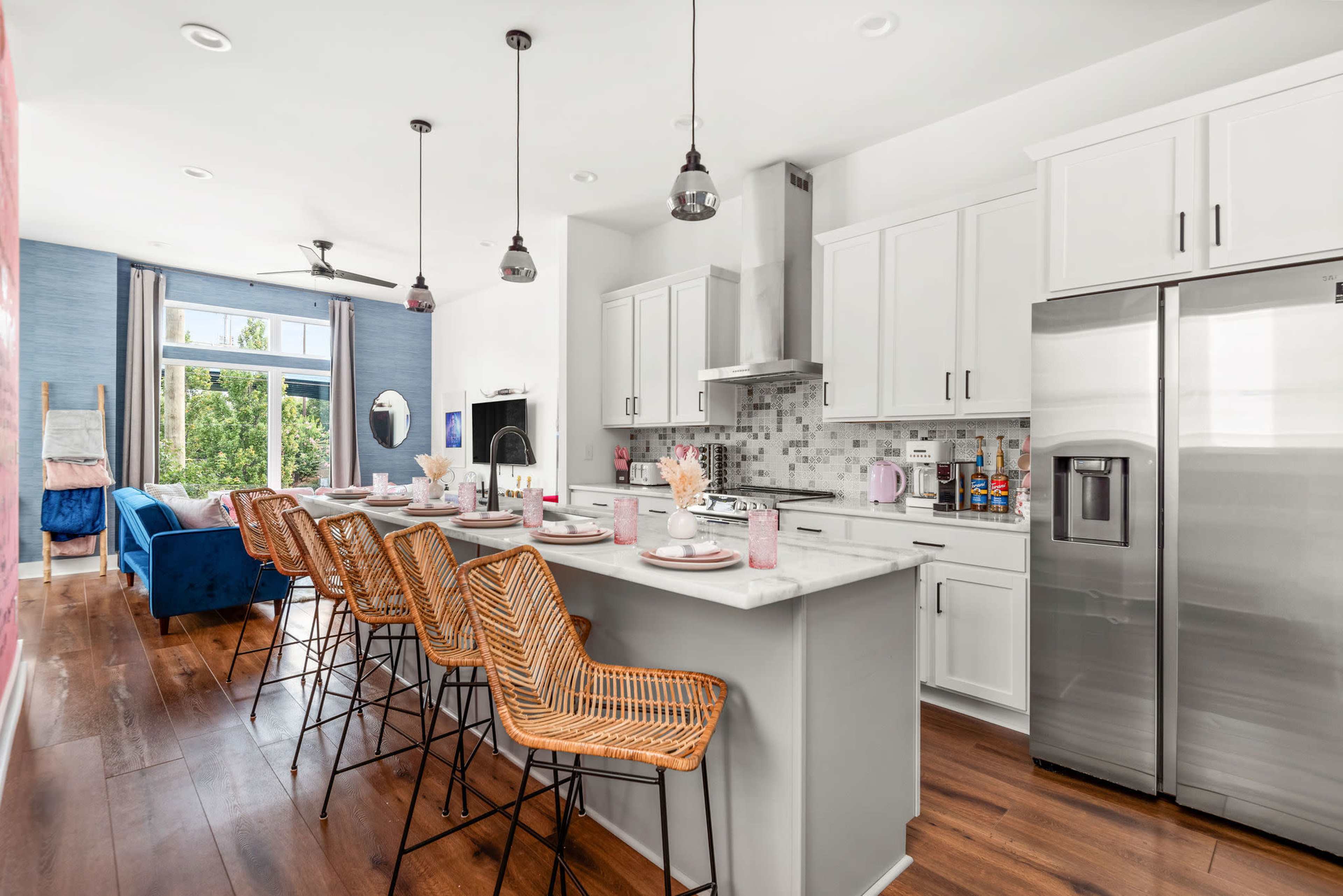 A modern kitchen with white cabinetry and a gray countertop features bar stools along a central island, with an adjacent living area visible in the background.
