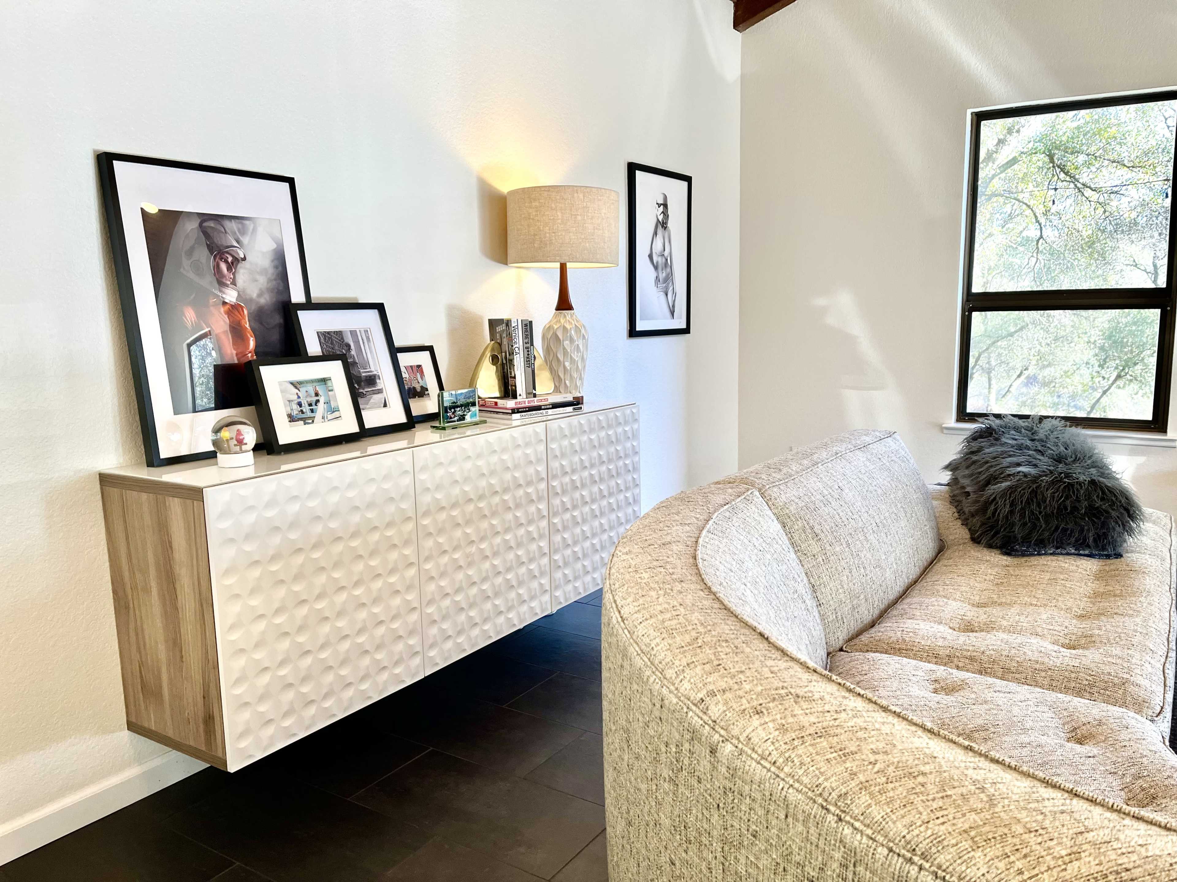 A textured white sideboard with framed photographs and a lamp is positioned next to a curved, beige sofa in a well-lit room with large windows.