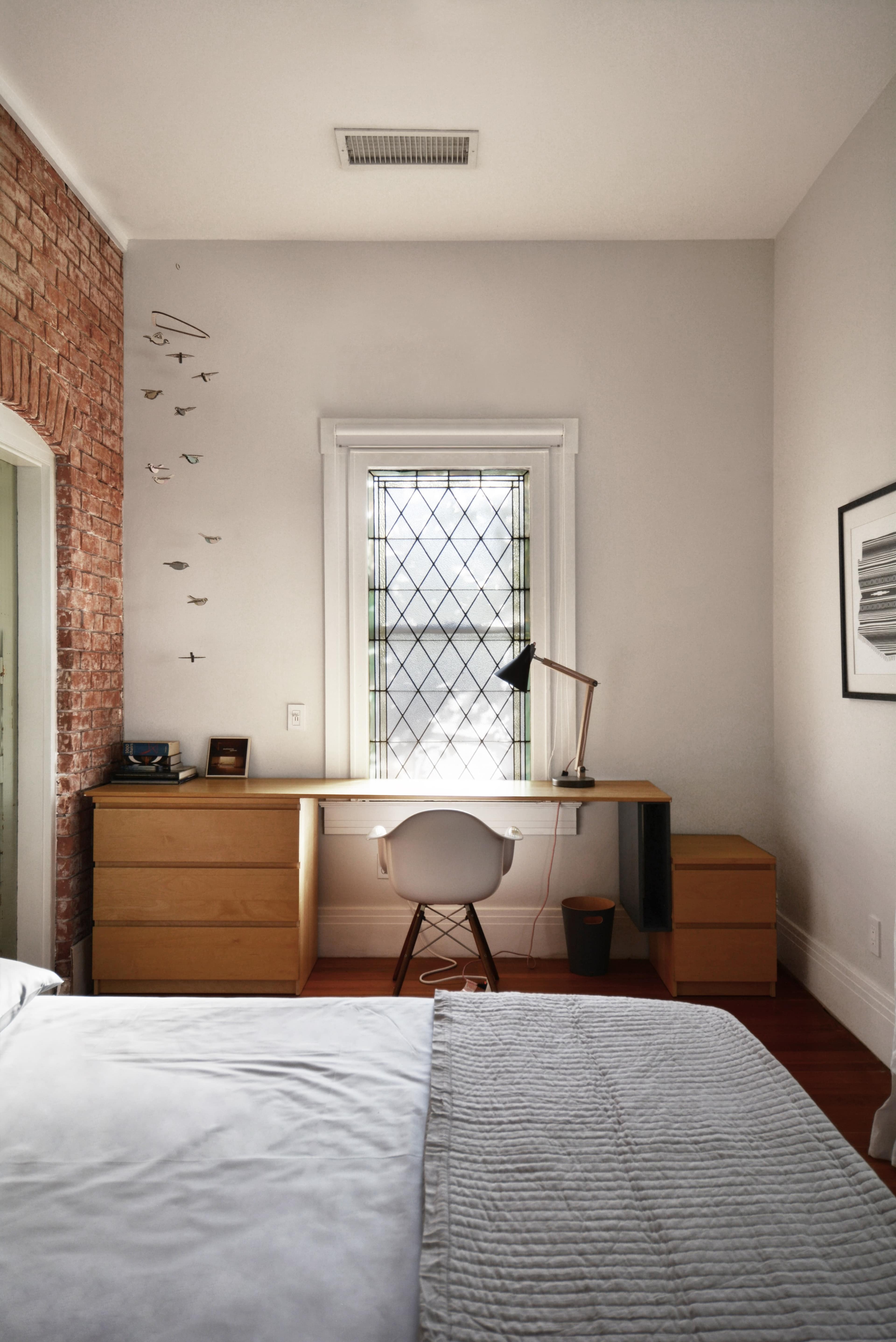 A minimalist bedroom featuring a wooden desk and chair next to a window with a diamond-patterned grid, complemented by a brick accent wall.