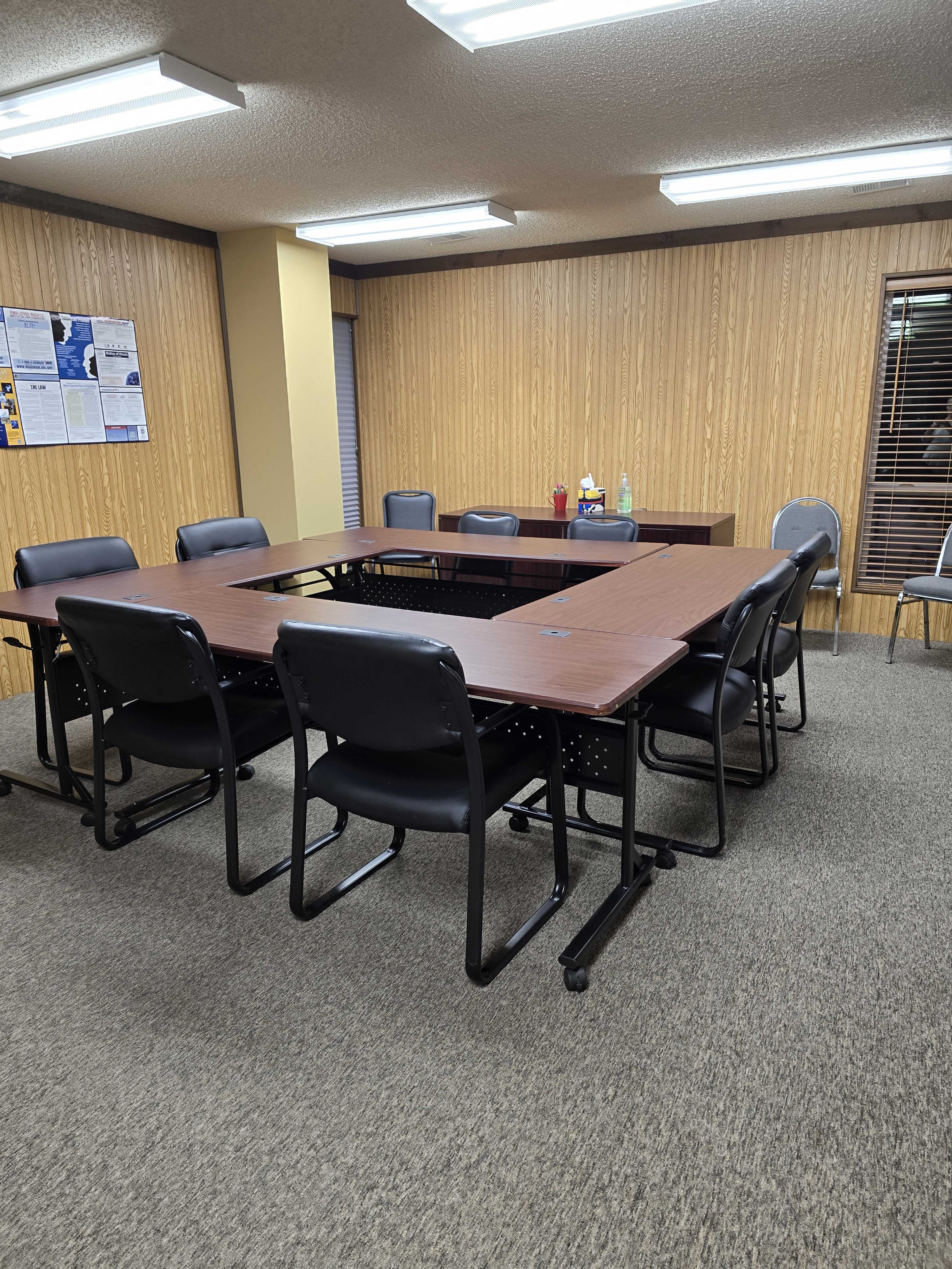 A large conference room features a central rectangular table surrounded by several black chairs, with wood-paneled walls and a small window.