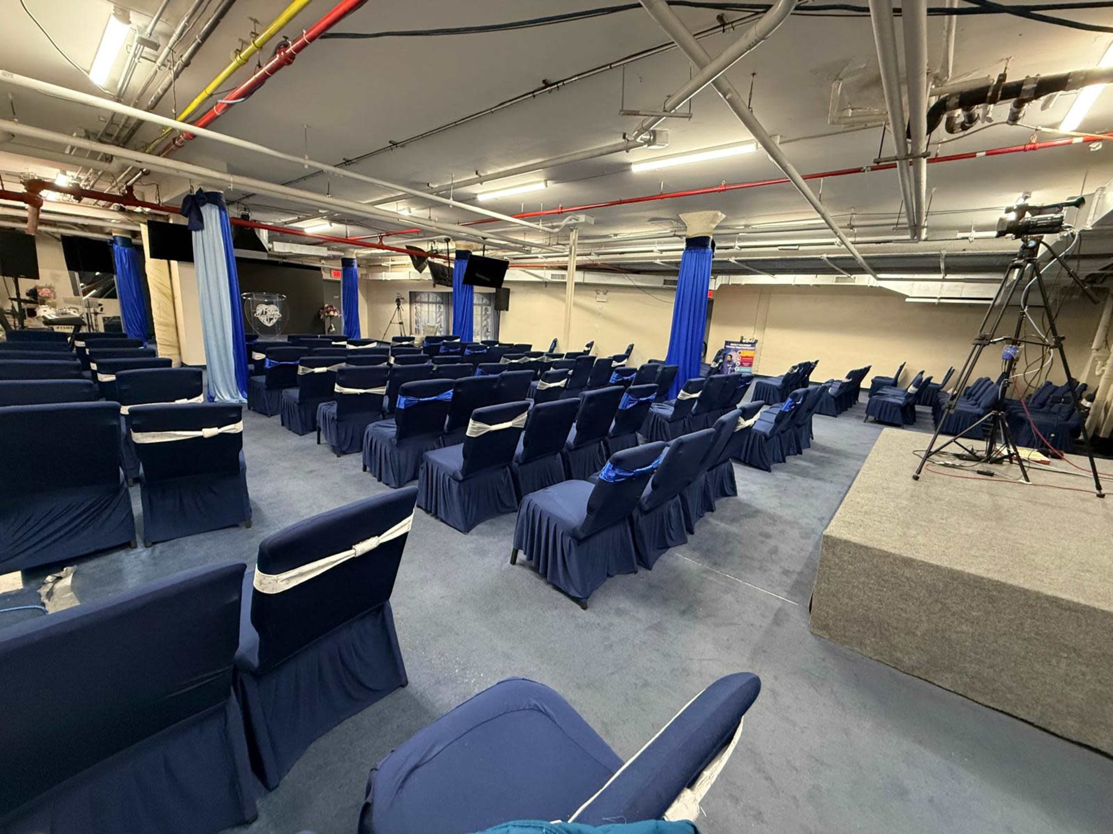 The image shows a large, empty conference room with rows of covered chairs and a raised platform for presentations.