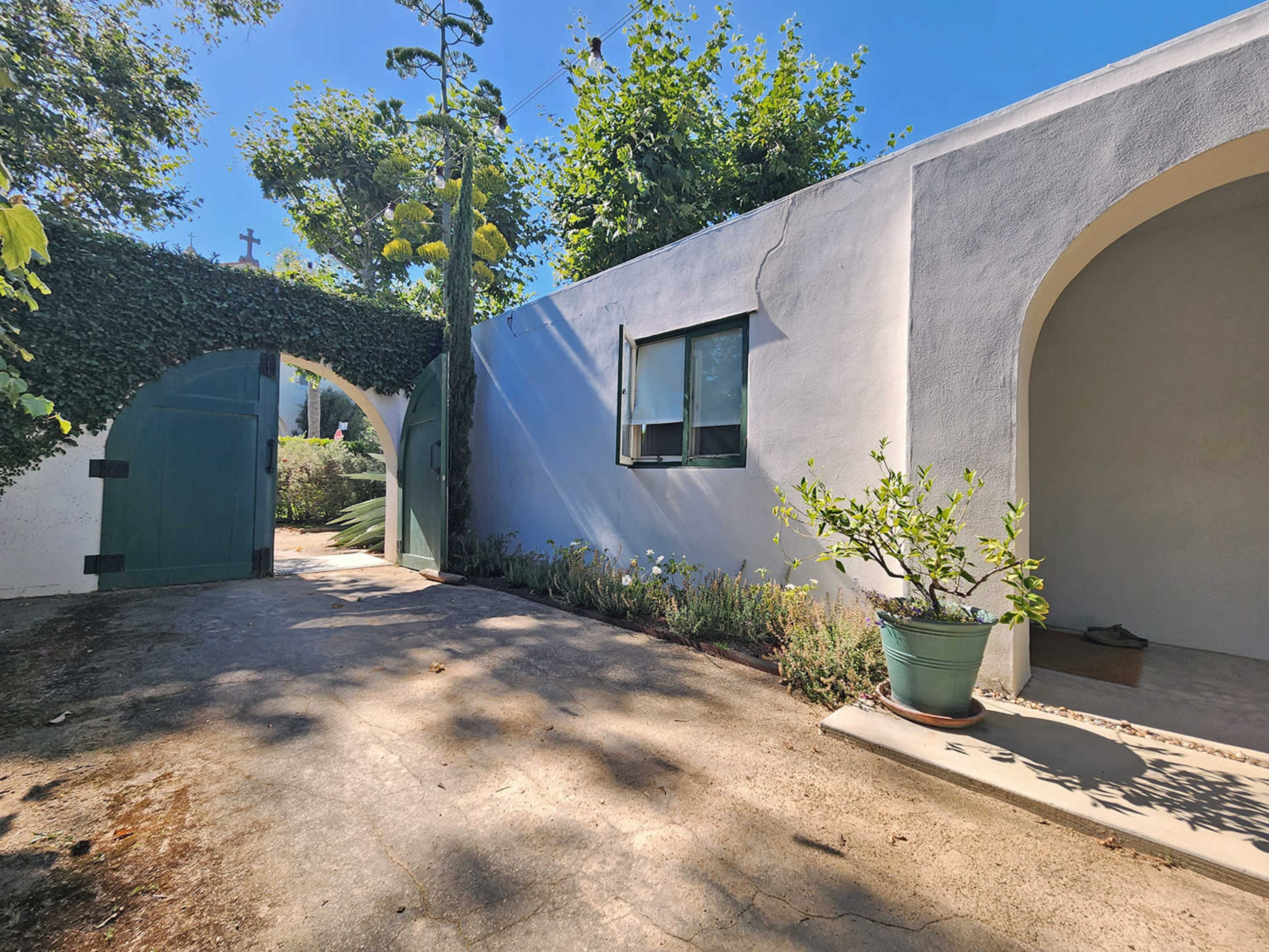 A pathway leads through a green gate to a white stucco building with a window and surrounding plants.