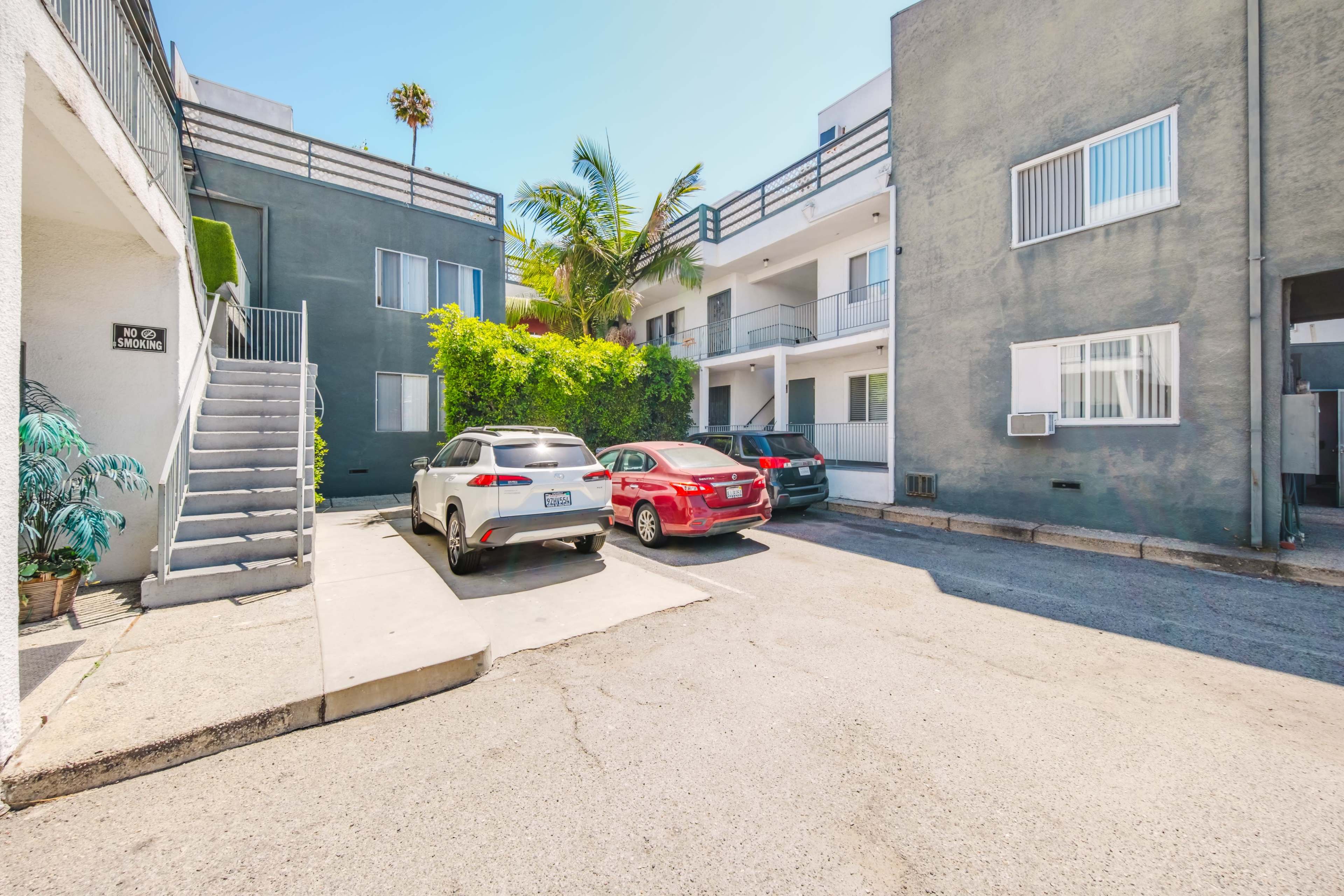 The image shows a multi-unit residential building with an adjacent parking area, featuring parked vehicles and palm trees in a sunny environment.