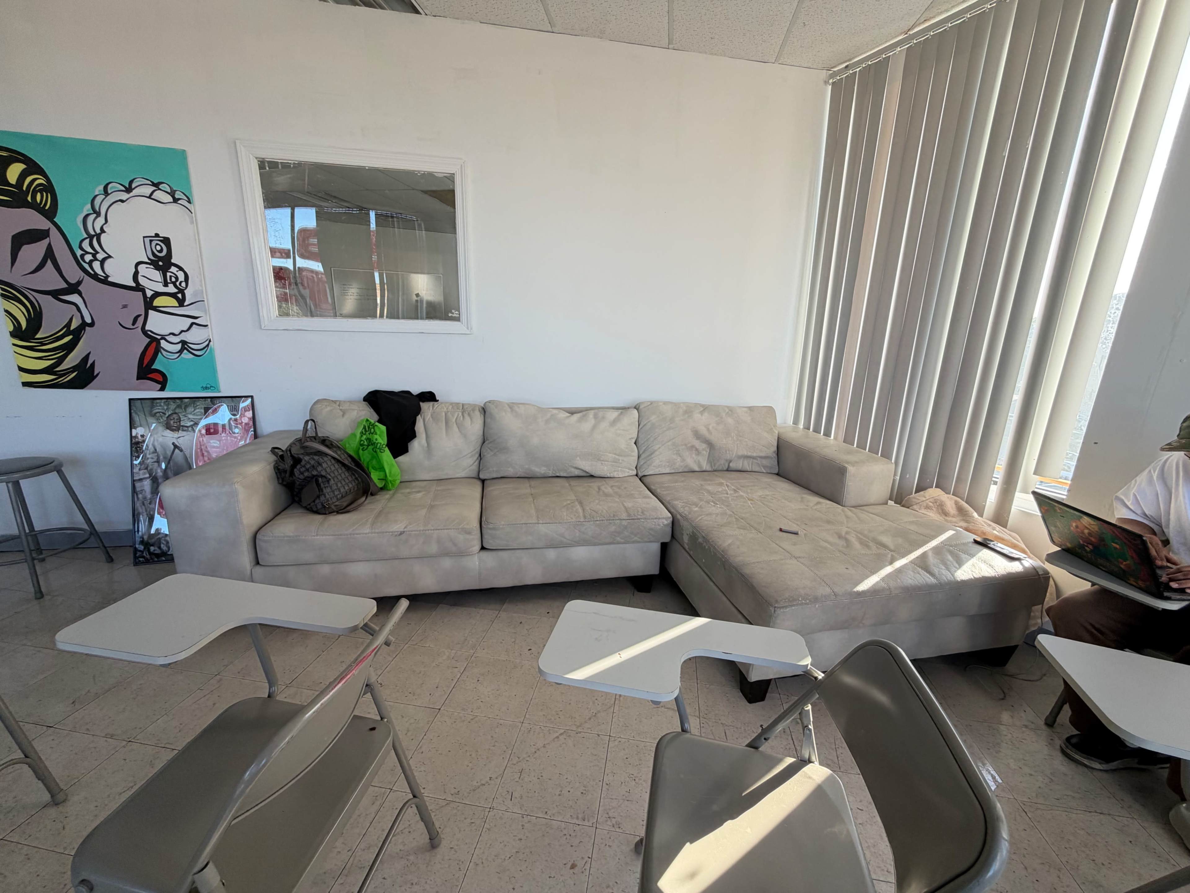 The image shows a light-colored sectional sofa in a room with vertical blinds, a window, and some empty desks.