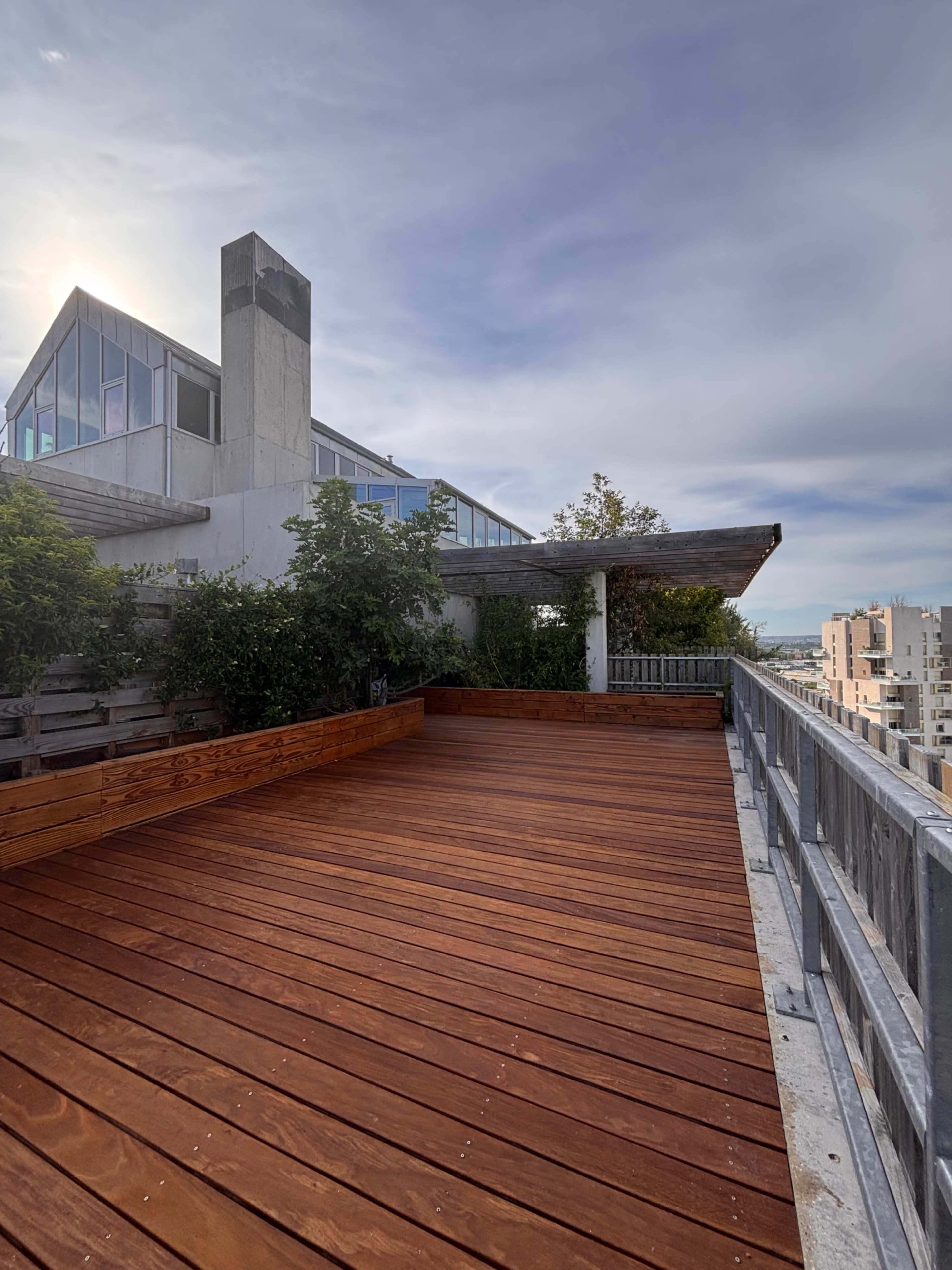 The image shows a rooftop terrace with wooden decking, surrounded by greenery and modern architecture in the background.