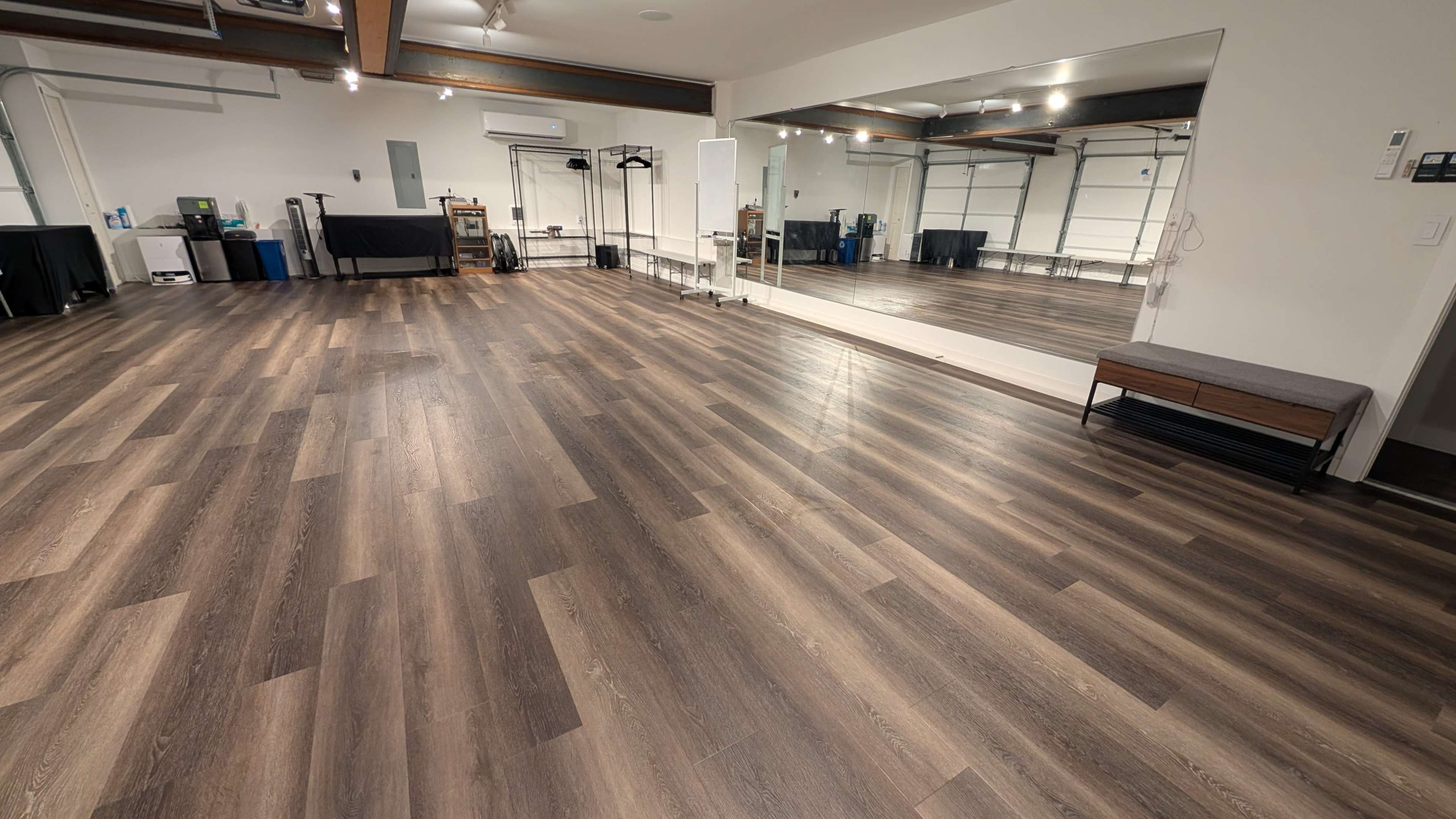 The image shows a spacious, empty indoor room with polished wooden flooring and a large mirror on one wall.