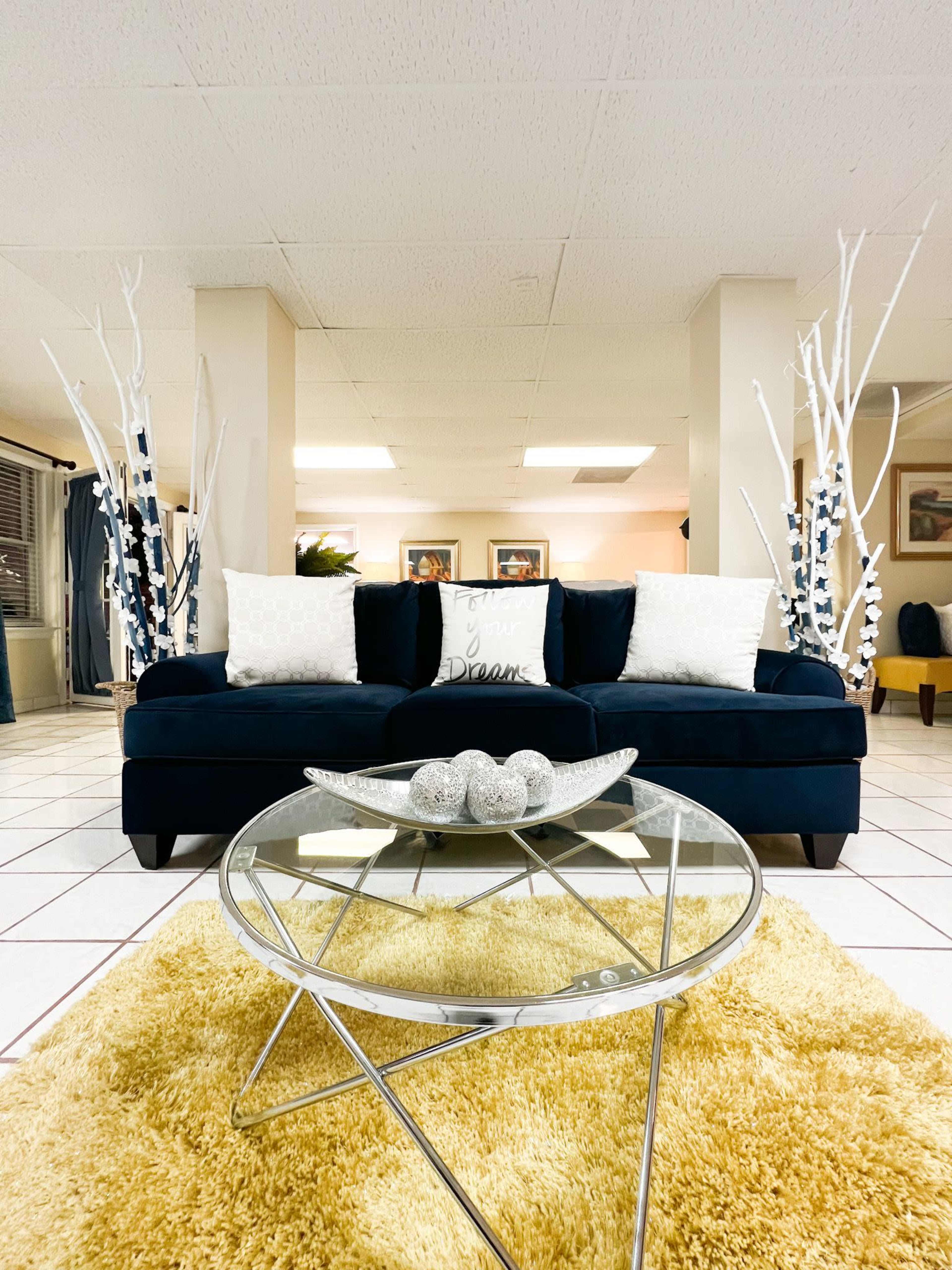 A blue sofa with decorative pillows sits against a light wall, accompanied by a glass coffee table and two white branch decorations on either side.