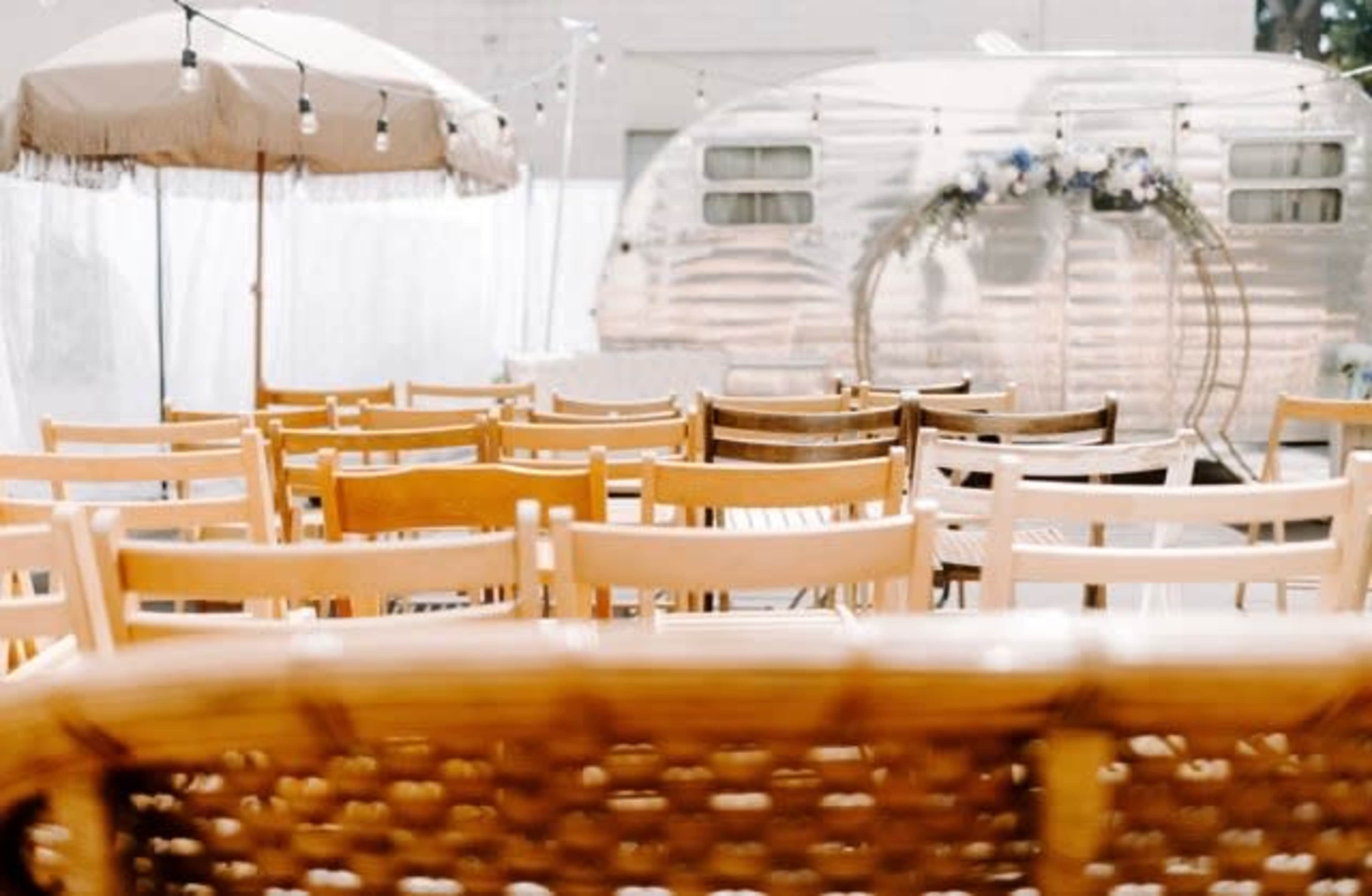 The image shows rows of wooden chairs arranged for an outdoor event, with an Airstream trailer and a shaded area in the background.