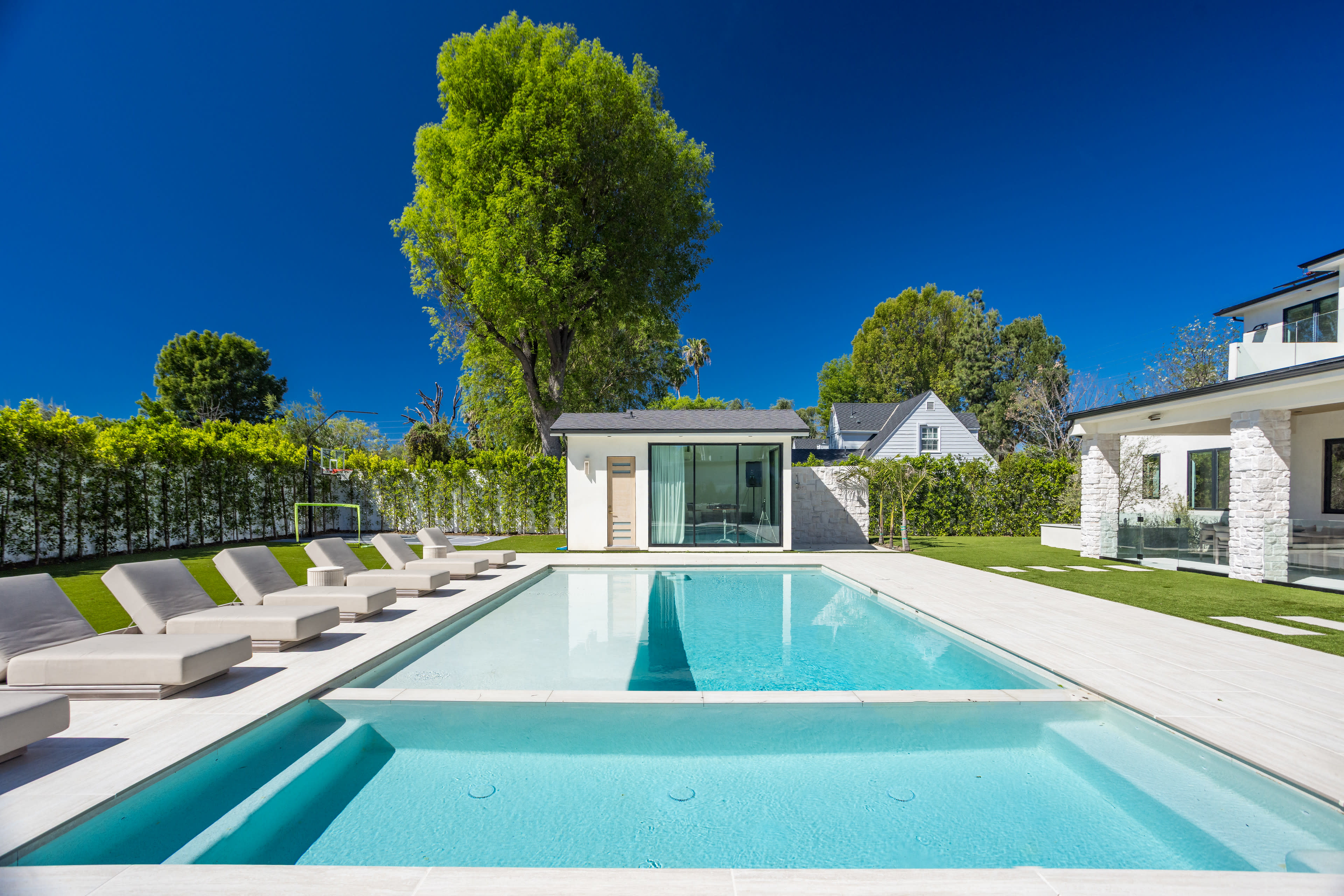 The image shows a modern outdoor swimming pool surrounded by lounge chairs and a landscaped yard with lush greenery.