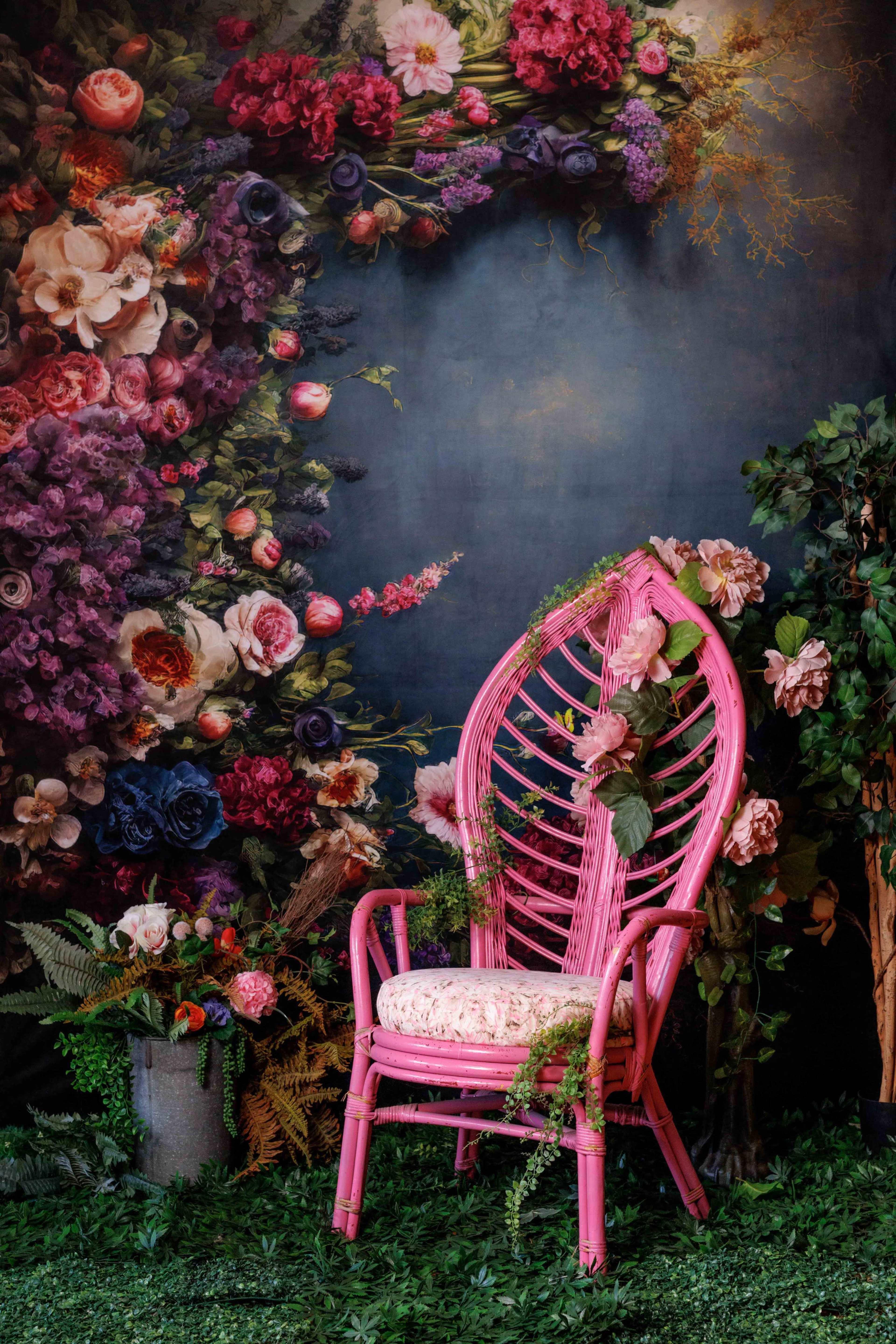 The image shows a pink wicker chair adorned with flowers and greenery, set against a dark floral backdrop.