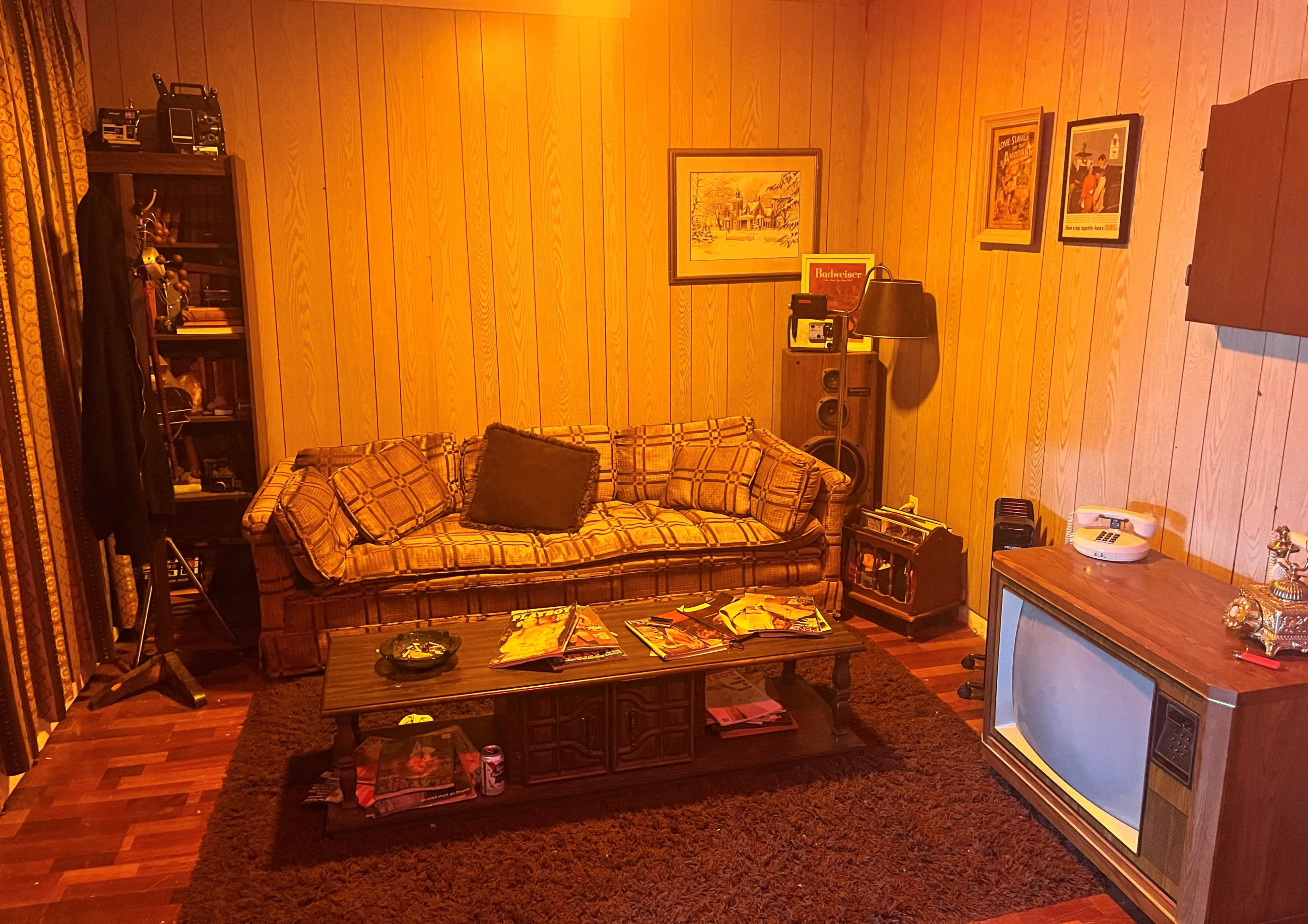 A vintage living room with a patterned sofa, a wooden coffee table displaying magazines, and an old television set against paneled walls.