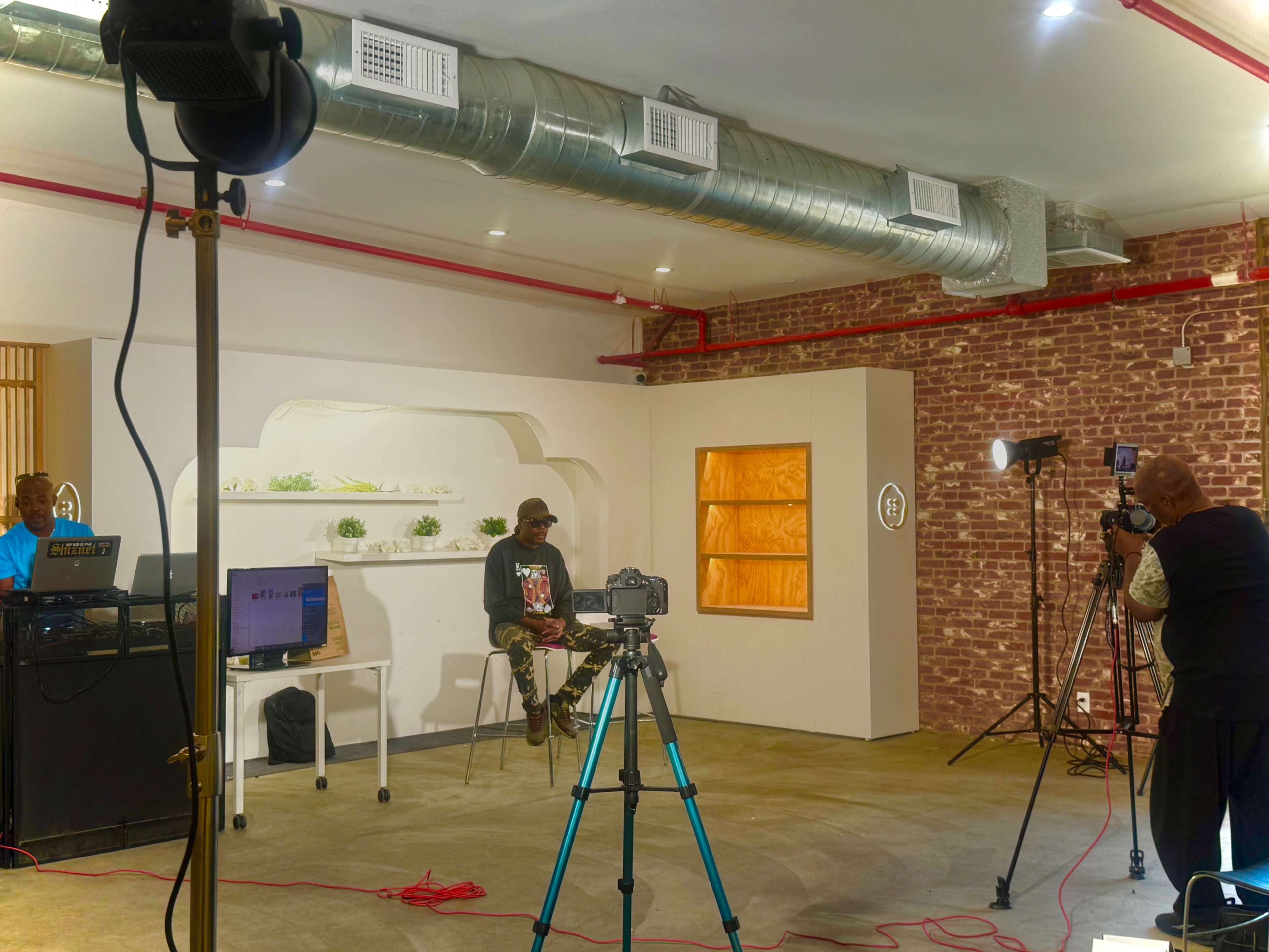 The image shows a recording studio setup with two photographers capturing a seated individual against a backdrop of a brick wall and decorative displays.