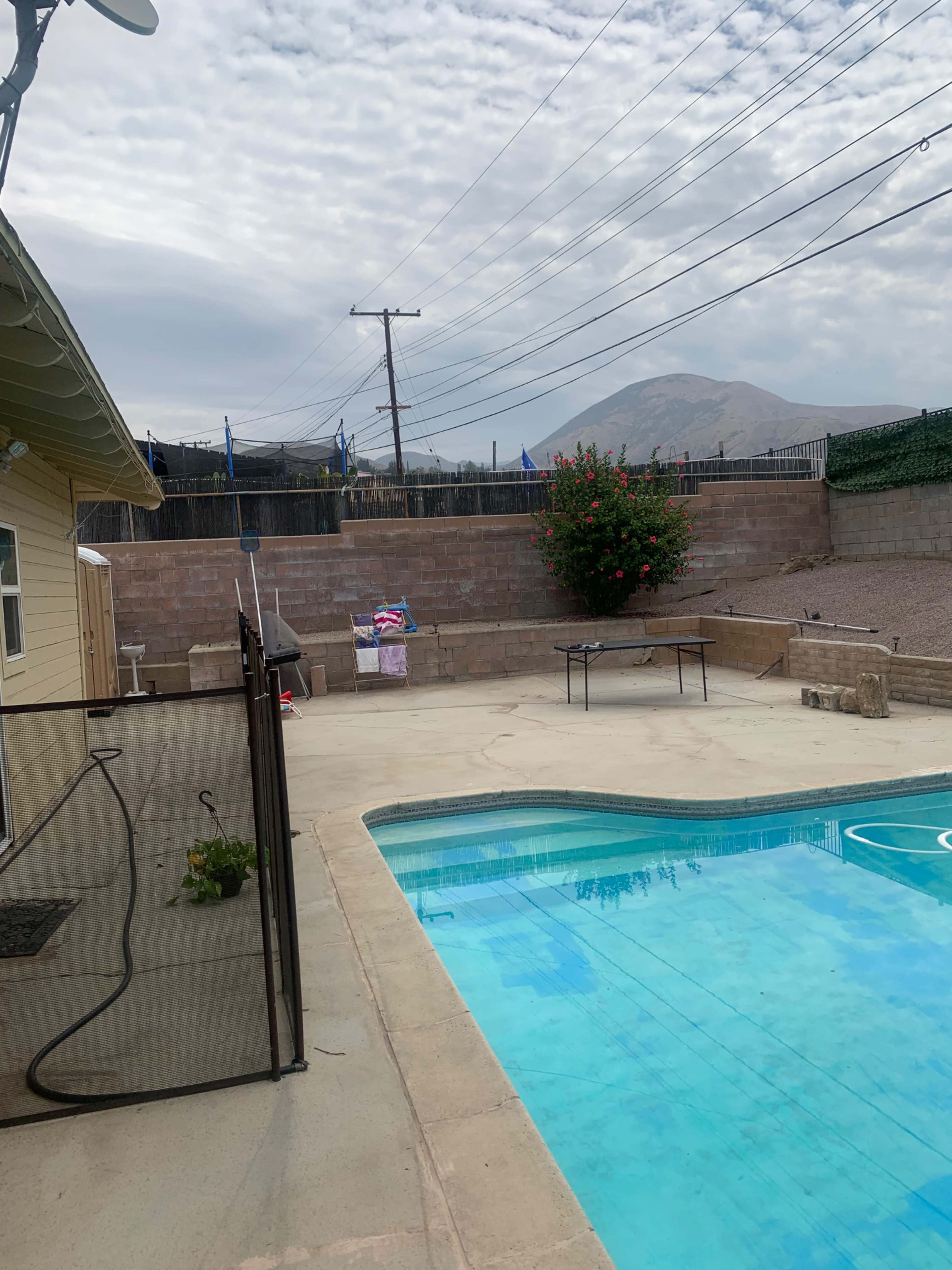 A backyard with a swimming pool, a patio area, and power lines in the background, surrounded by a low wall and a landscaped hill.